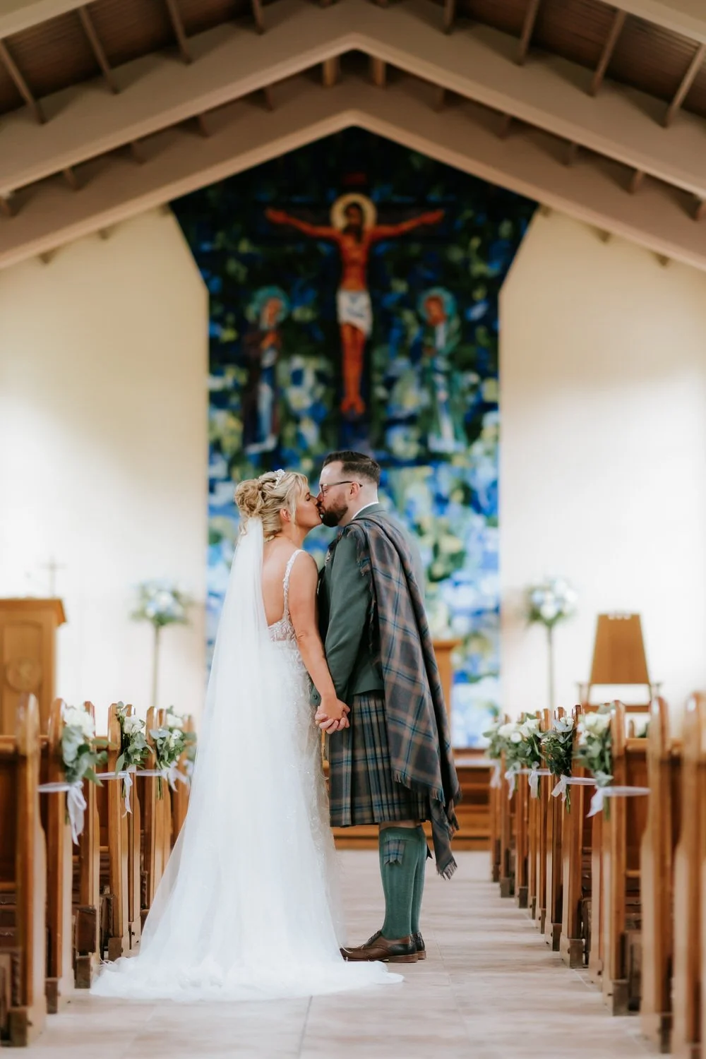 Bride and groom kissing inside a church, holding hands, with a large colorful cross painting behind them and decorated pews with white flowers.