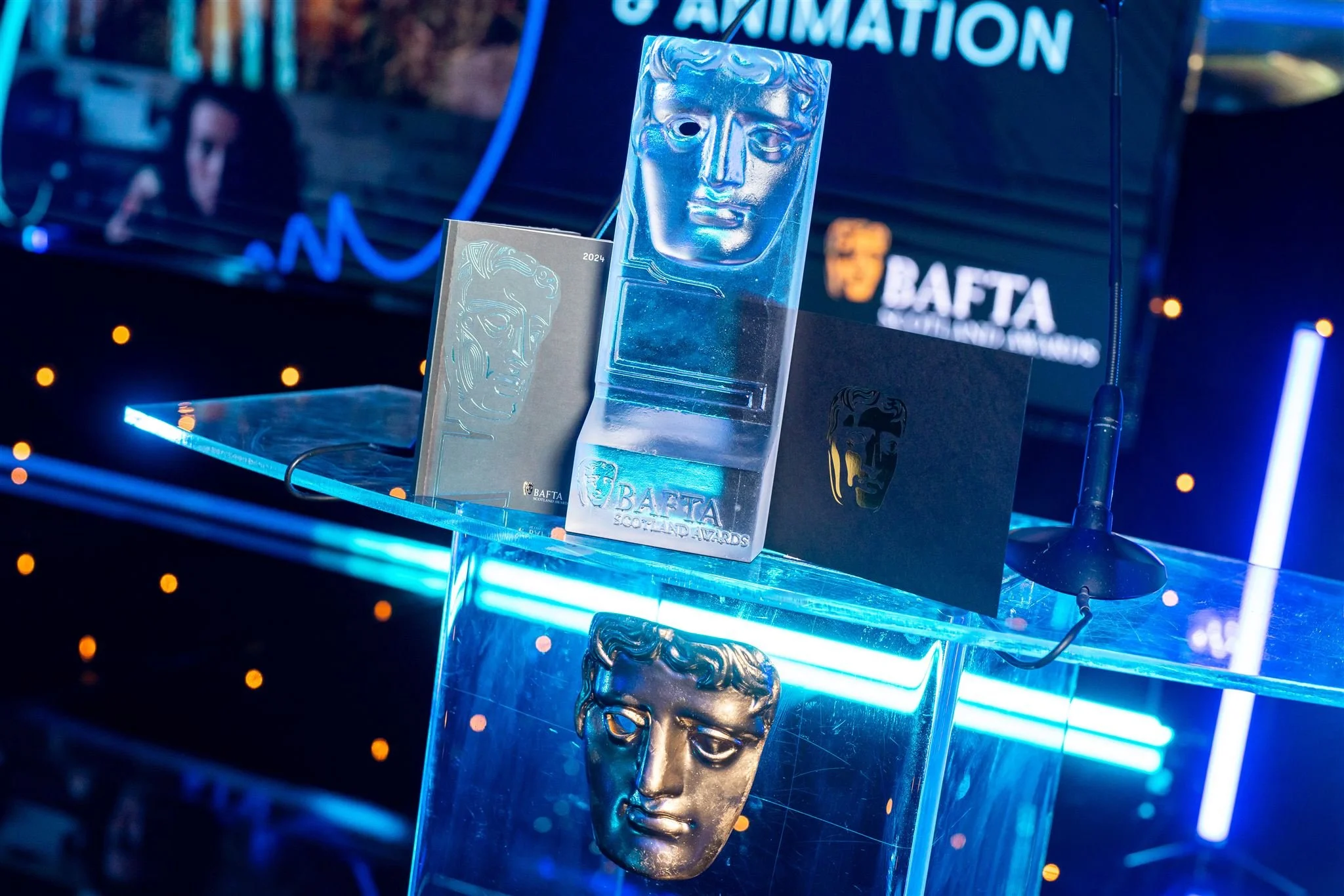 BAFTA awards trophy and plaques displayed on a transparent table illuminated by blue neon lights at an event.