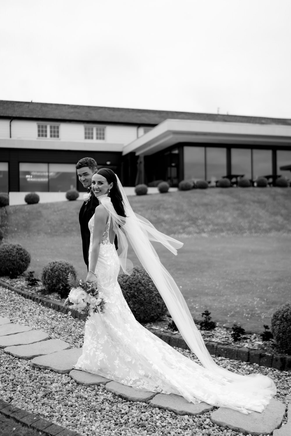 Black and white photo of a bride and groom walking on a stone path outside a modern building with large glass windows, smiling and holding a bouquet of flowers.
