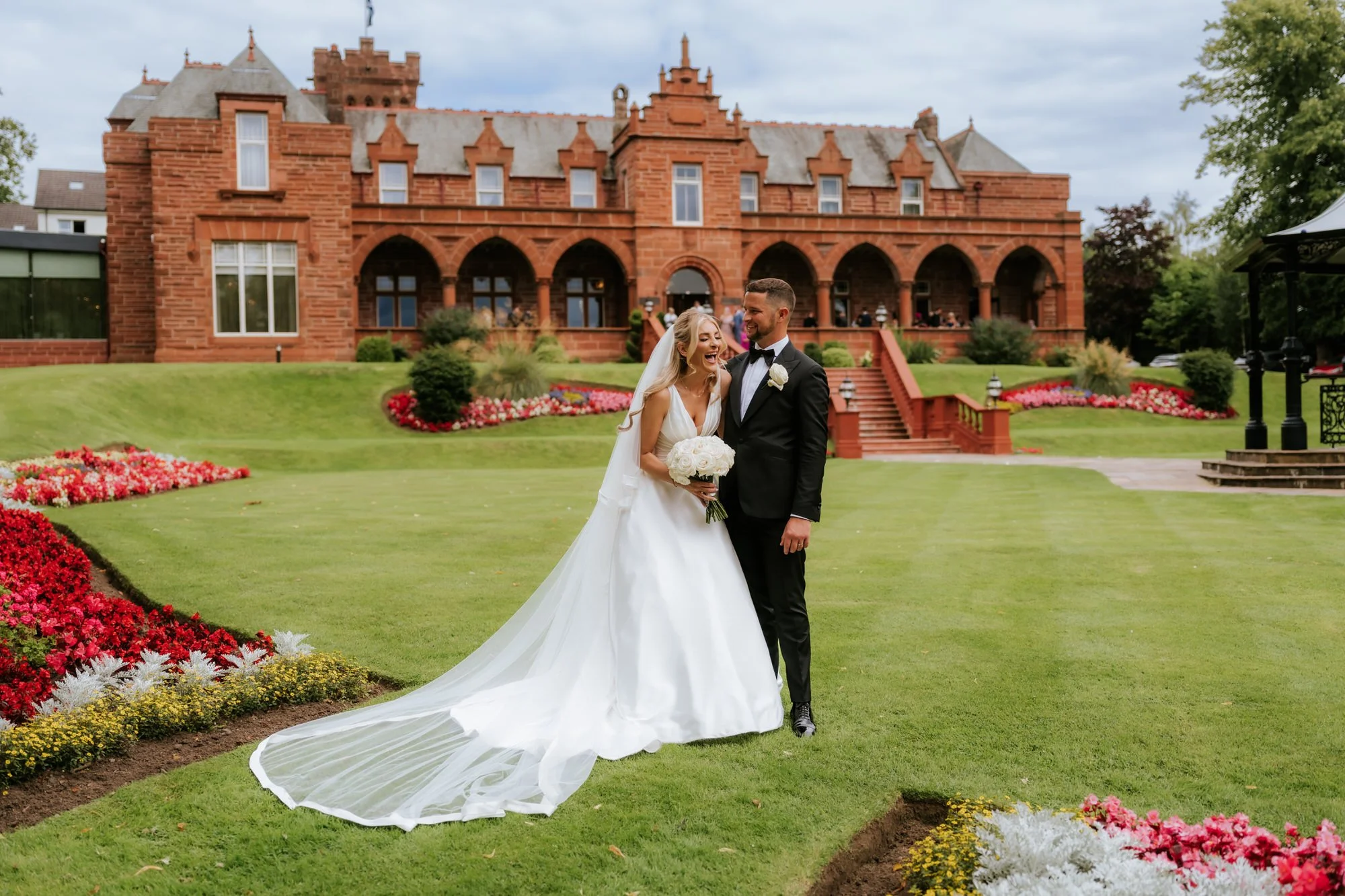 A bride and groom standing on a well-manicured lawn in front of a large red brick historic mansion. The bride wears a white wedding gown and veil, holding a bouquet of white roses, while the groom is dressed in a black tuxedo with a bow tie. They are