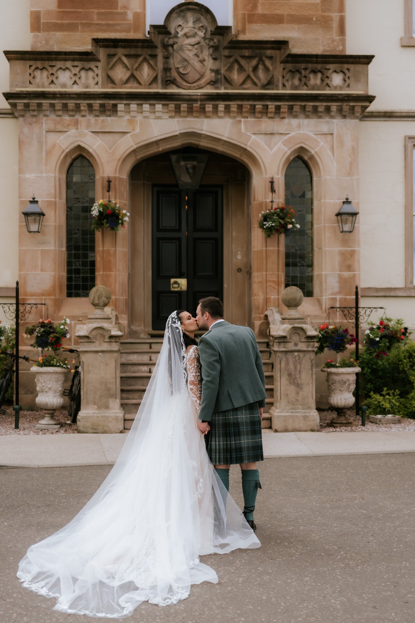 Bride and groom kissing in front of a historic building with stairs, black door, and flower arrangements.
