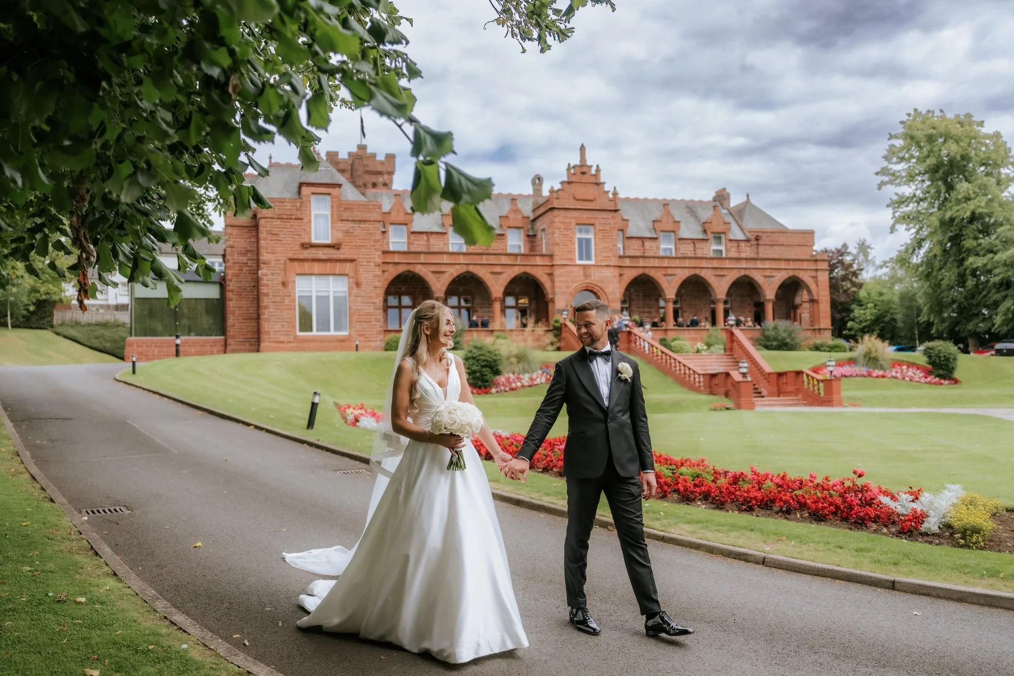 A bride and groom walking hand in hand down a paved path outside a red-brick building, surrounded by well-manicured grass and colorful flower beds, during their wedding celebration.