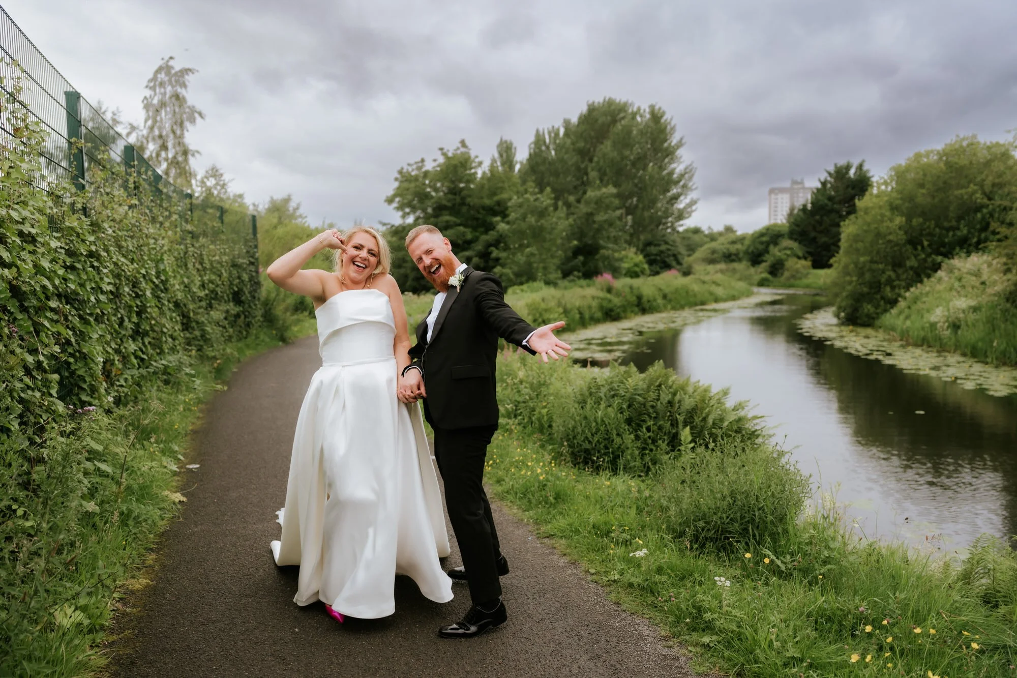 A newlywed couple in wedding attire standing on a paved path by a river, surrounded by greenery and trees, under a cloudy sky, smiling and posing playfully.
