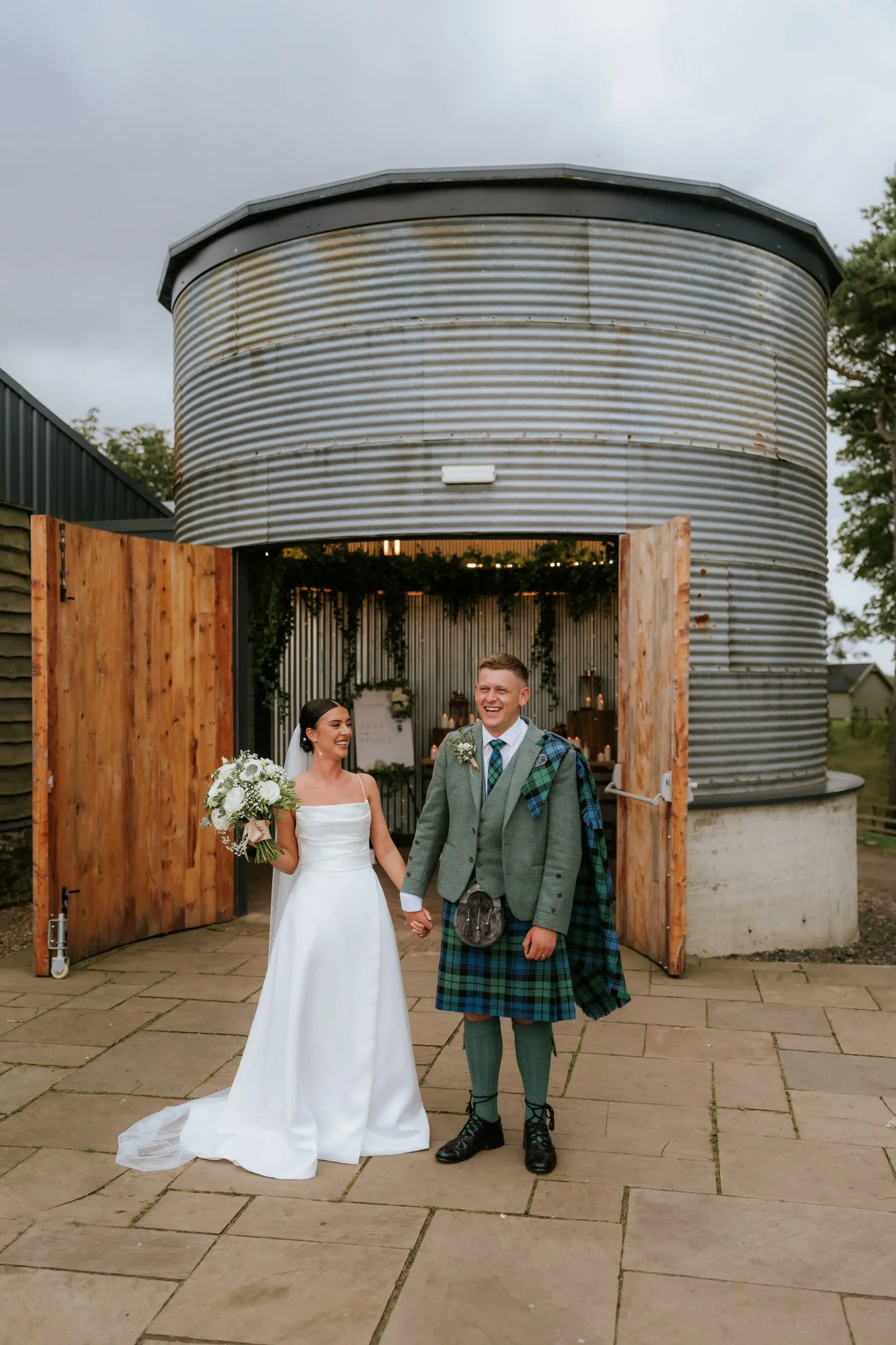 A bride and groom holding hands and smiling outside a barn at their wedding celebration. The bride is wearing a white dress and holding a bouquet, while the groom is dressed in a kilt and jacket, with a tartan sash and a sporran. The barn has large w