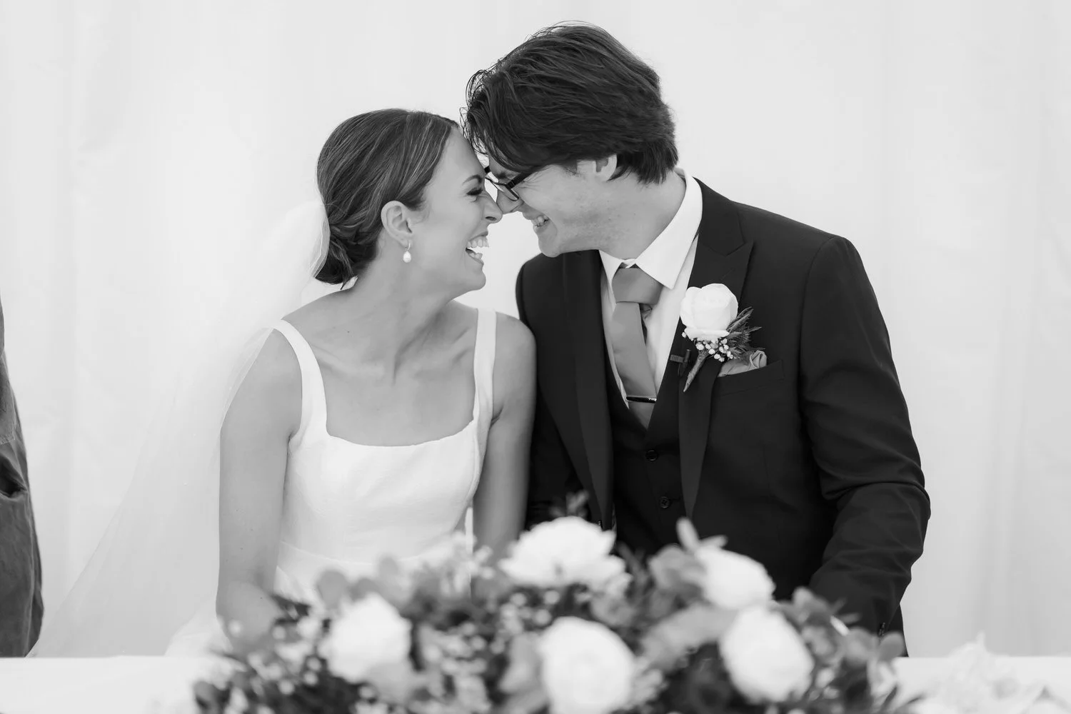 Black-and-white photo of a bride and groom smiling and touching foreheads at their wedding reception, with a floral arrangement in the foreground.