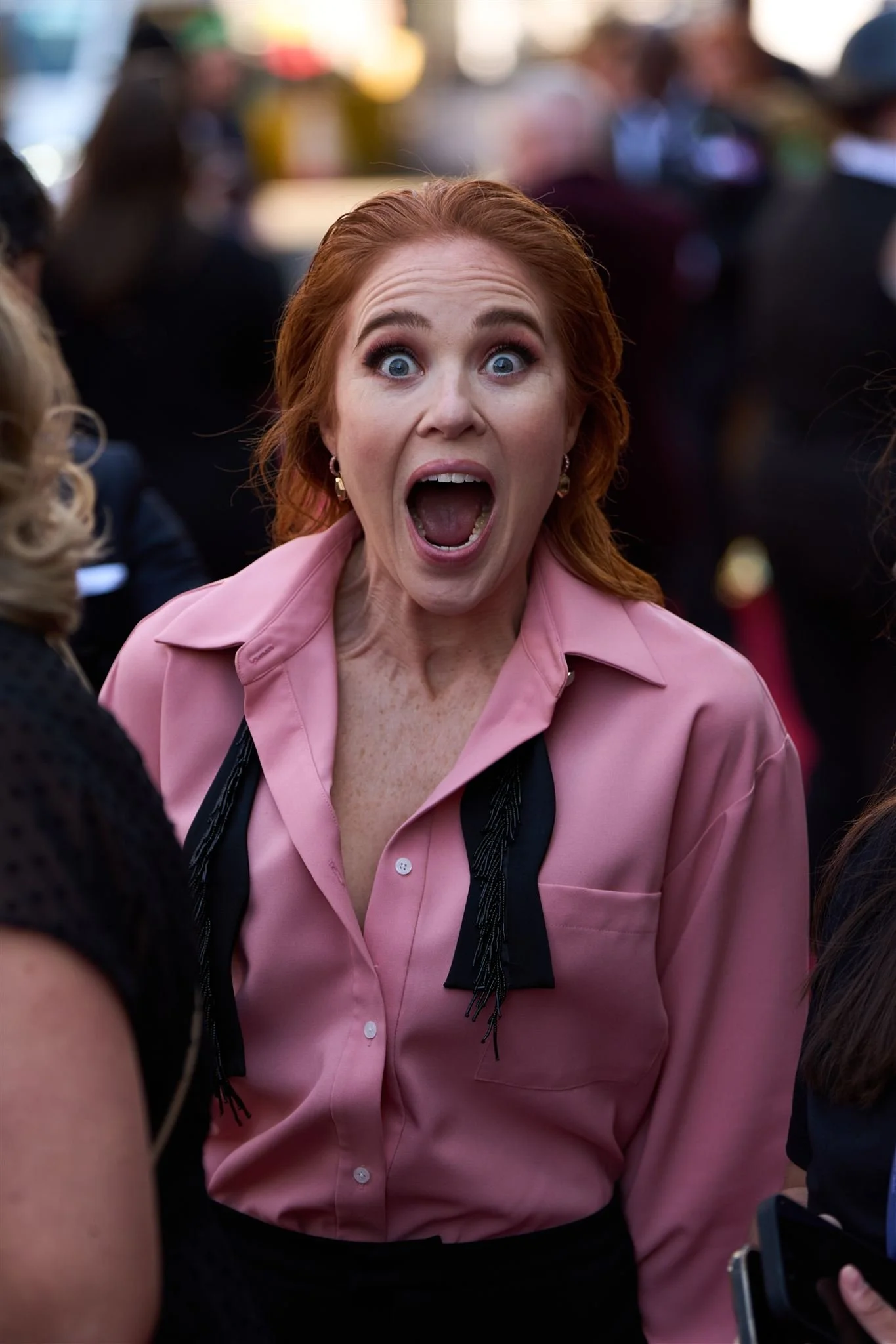 A woman with red hair wearing a pink shirt and black necktie, with an expression of shock or surprise, mouth wide open, in a crowd of people.