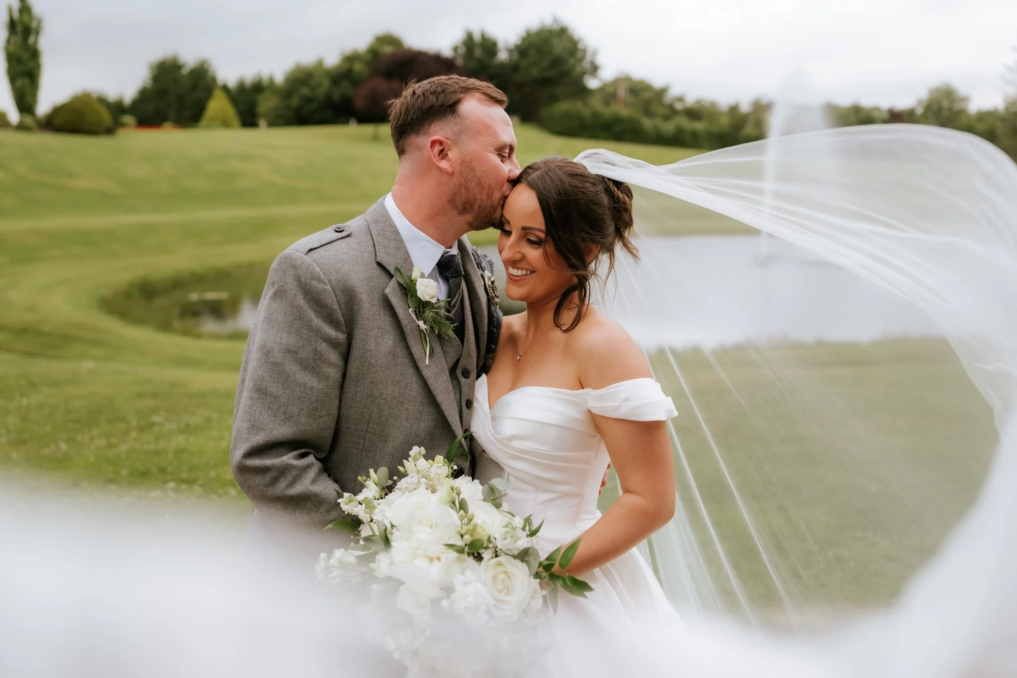 A newlywed couple on their wedding day outside, with the groom kissing the bride on the forehead as she smiles. The bride is wearing a white off-the-shoulder wedding dress and holding a bouquet of white flowers. The groom is dressed in a gray suit wi