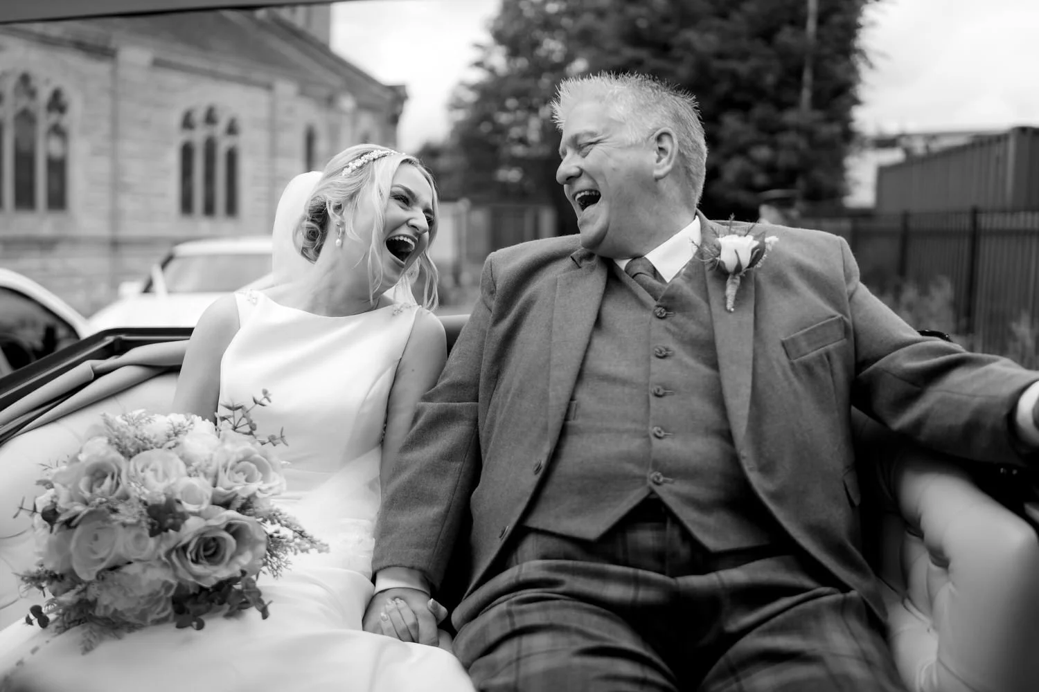 A bride and an older man, possibly her father, share a joyful laugh while sitting in a wedding car. The bride holds a bouquet of flowers and wears a wedding dress and veil, while the man wears a suit with a boutonniere. They are outdoors with a build