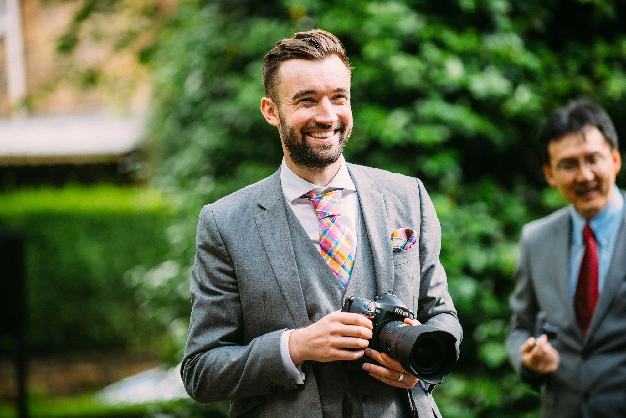 A smiling man in a gray suit holding a Nikon camera outdoors, with a man in a gray suit and red tie in the background, and greenery behind them.
