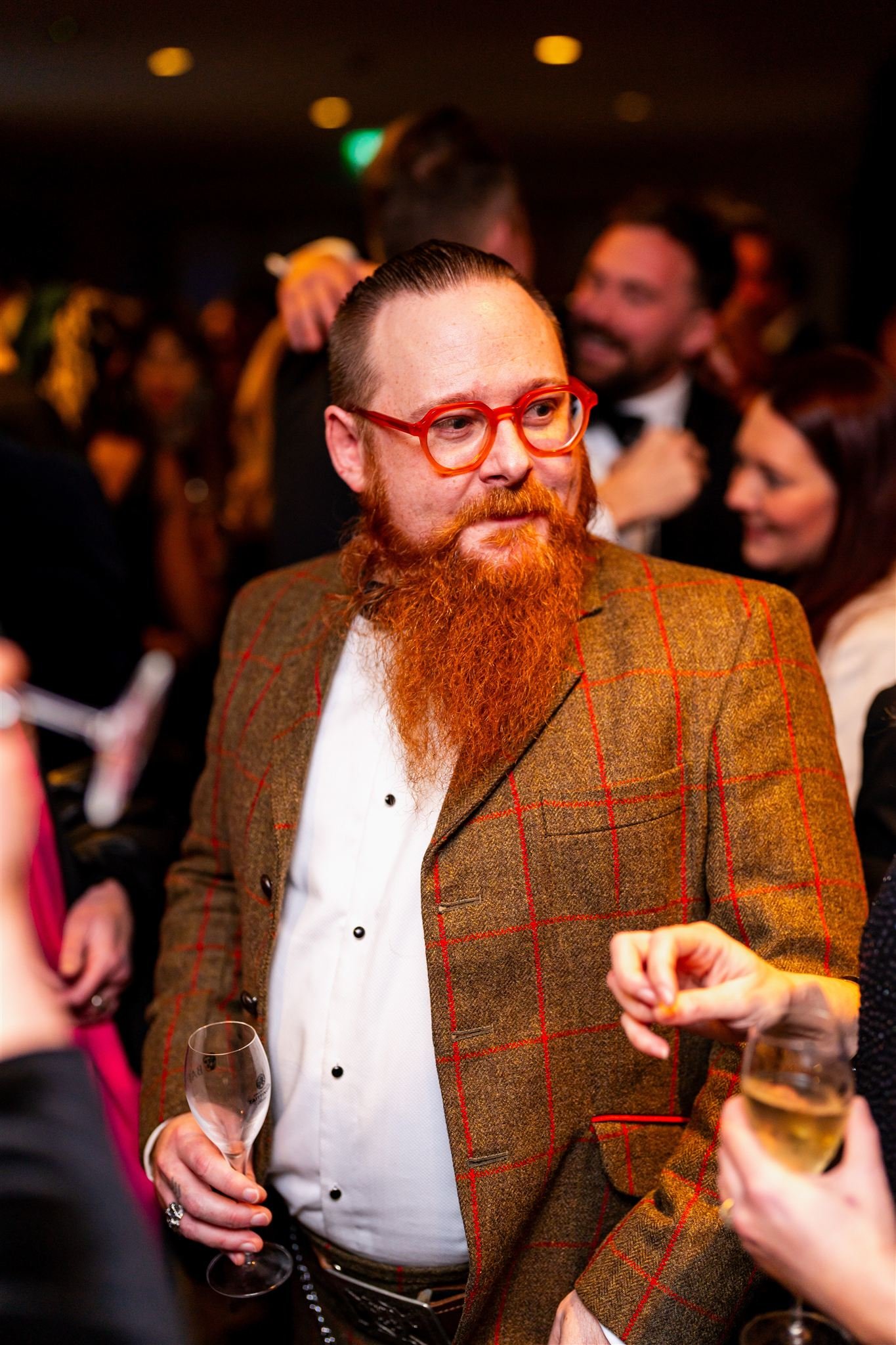 Man with red hair, beard, glasses, wearing a brown checked blazer and white shirt, holding a glass of wine at a social event.