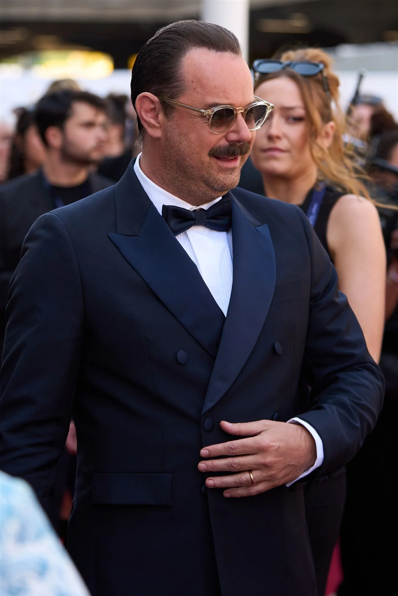 Man in a tuxedo with glasses, bowtie, and a wedding ring, standing among a group of people at an outdoor event.