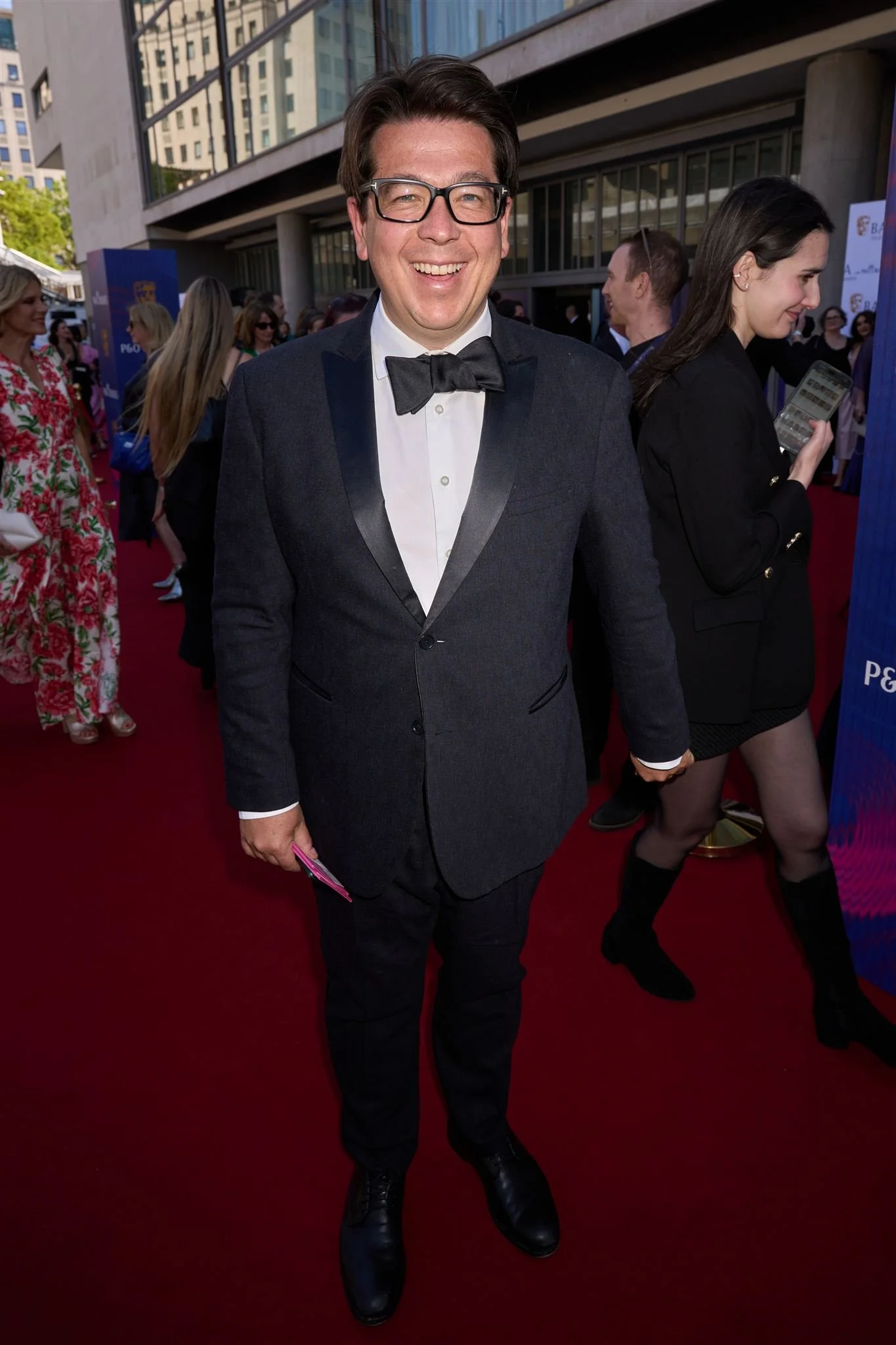 A man in a tuxedo with glasses, smiling on a red carpet at an event, with people and a cityscape in the background.