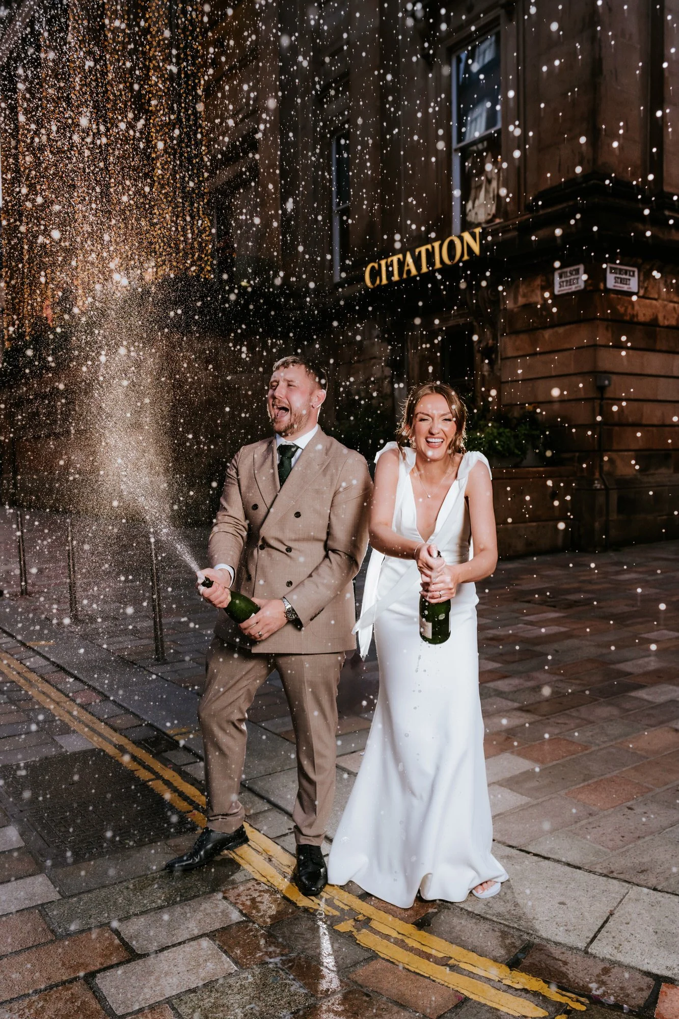 A newlywed couple celebrating outside at night, spraying champagne, with fireworks and sparks in the air. The groom wears a beige suit and tie, and the bride wears a white dress. They are standing on a city street near a building with a sign that rea