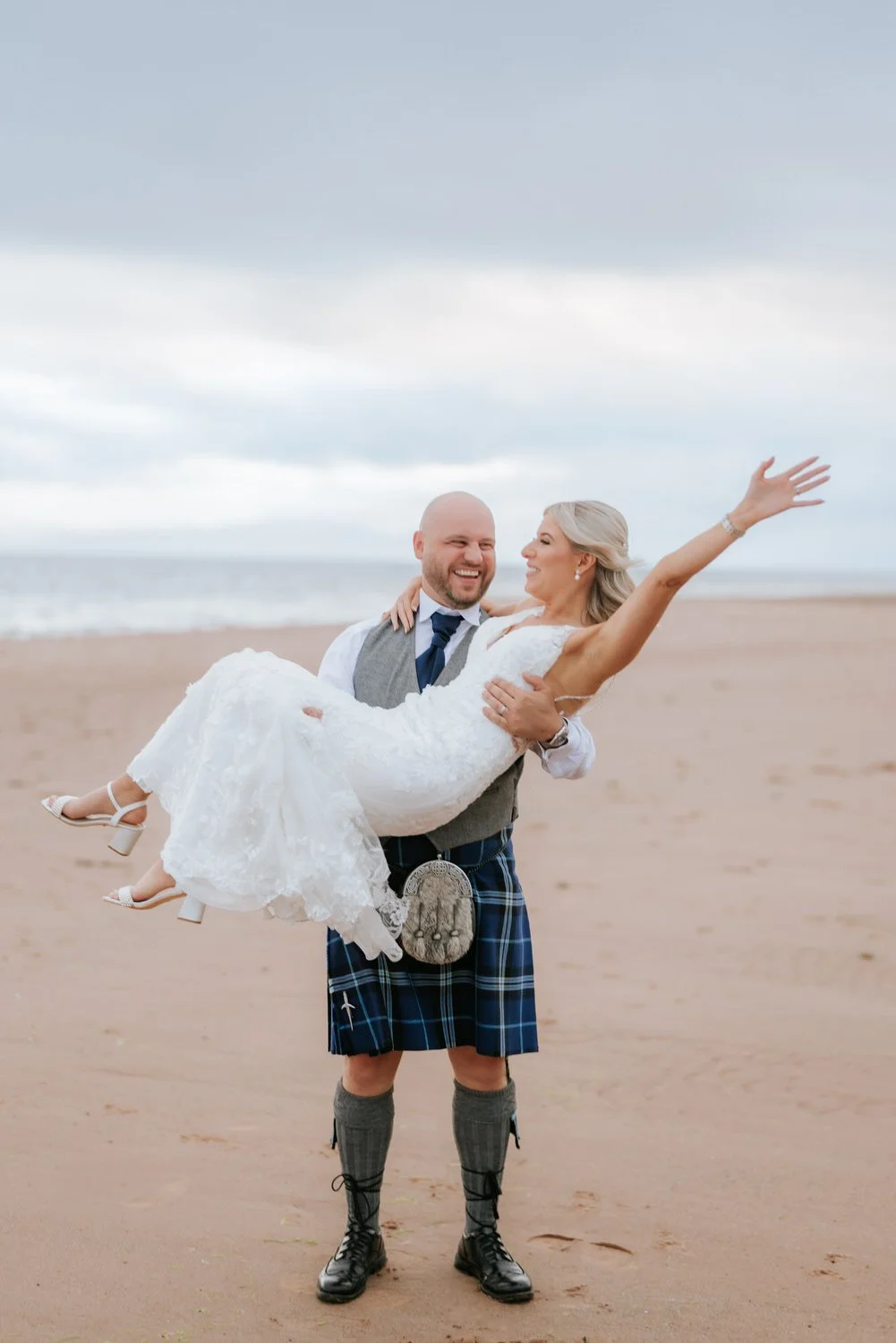 A man in a kilt holding a woman in a wedding dress on a beach, with the woman smiling and extending her arm.