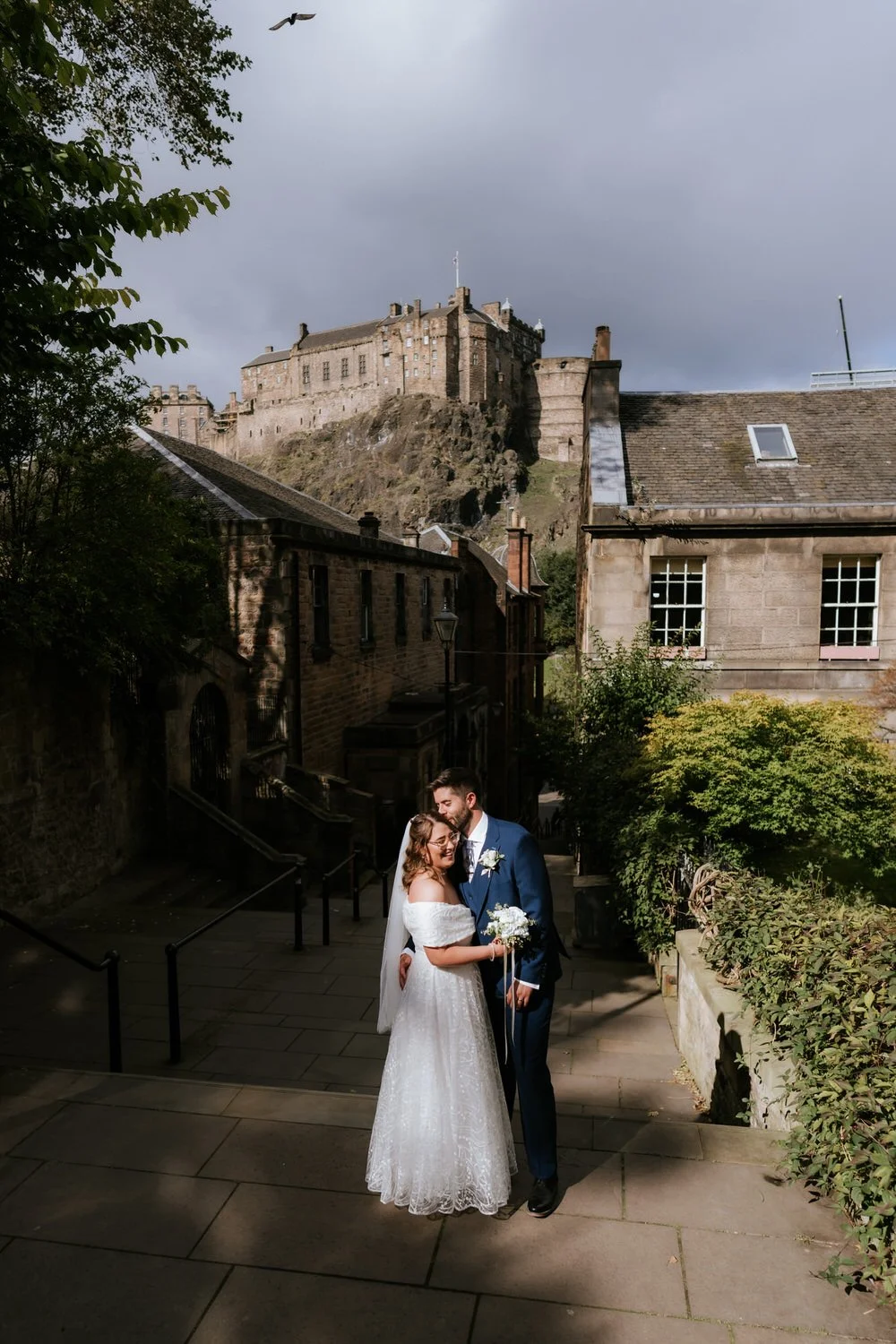 A bride and groom on their wedding day in an outdoor setting with a historic castle on a hill in the background. The bride is wearing a white off-the-shoulder wedding dress and holding a bouquet, while the groom is in a navy suit. They are smiling an