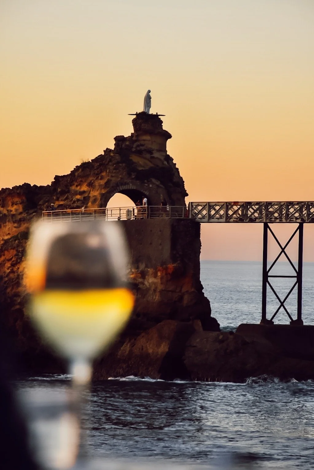 Une roche en mer avec une statue au sommet et un pont métallique, vue pendant le coucher du soleil. Un verre flou au premier plan.