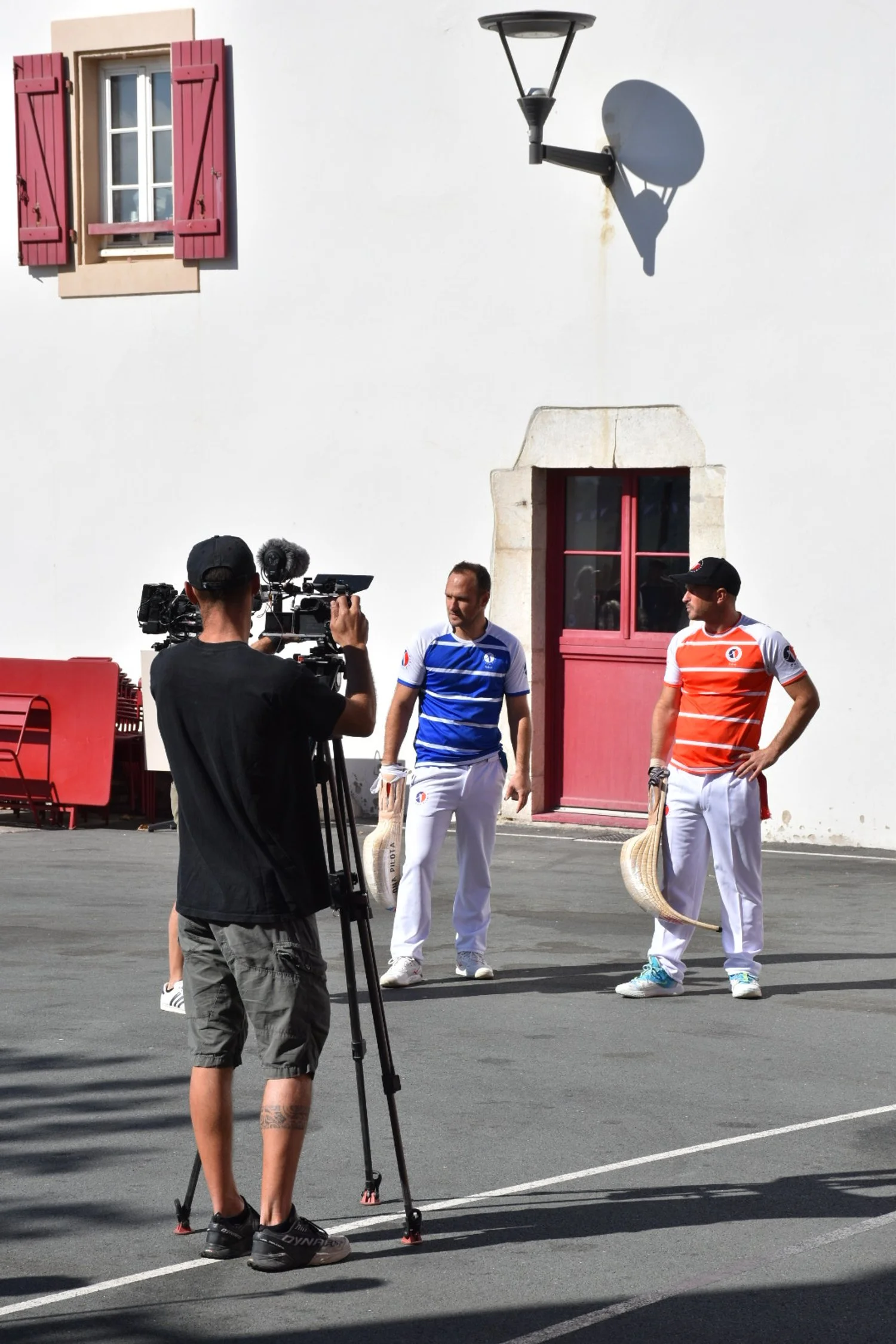Deux joueurs de pelote basque se préparent pour un match. L'un porte un maillot bleu, l'autre un maillot orange. Un cameraman les filme avec une caméra sur trépied devant un mur blanc.