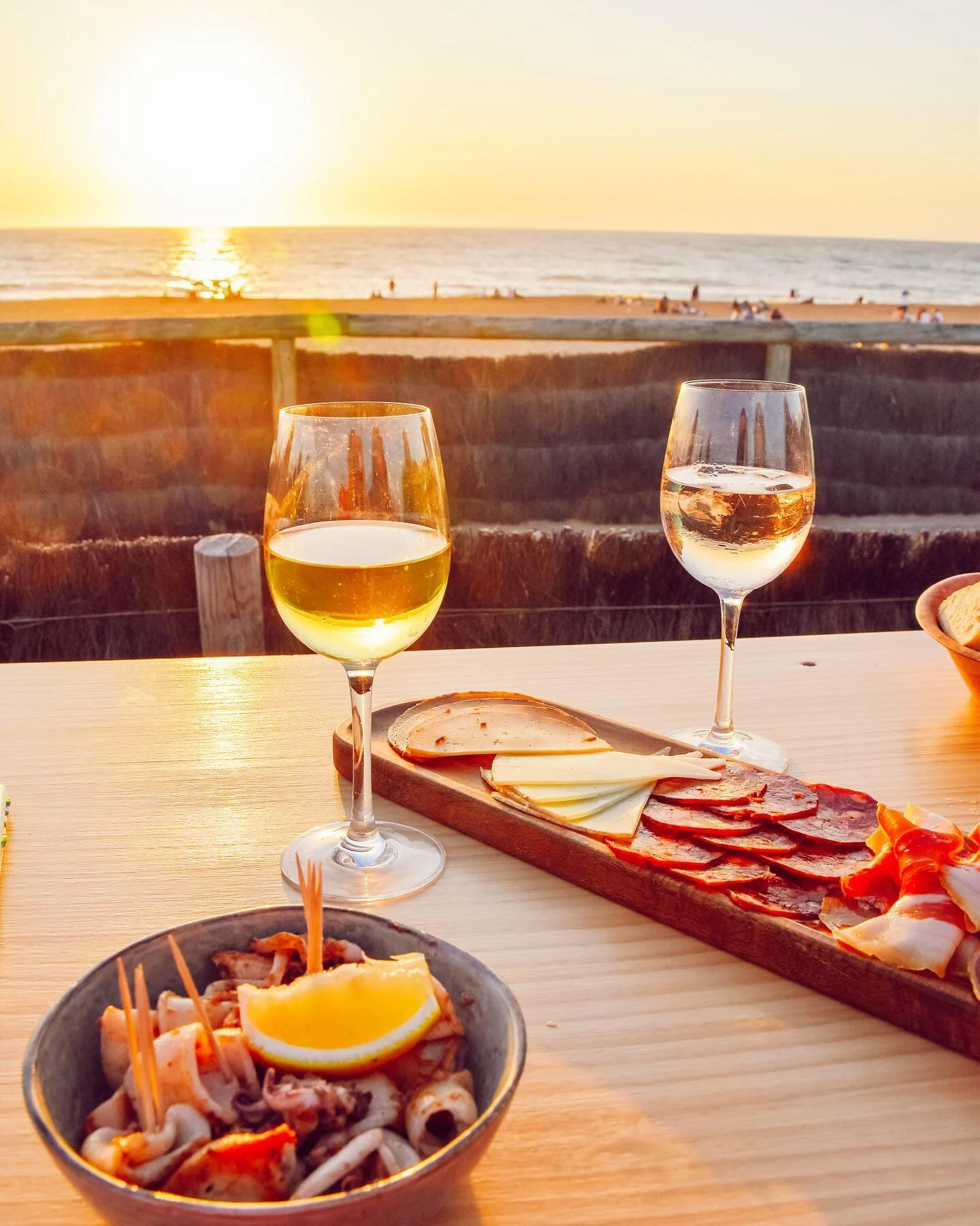 Table avec deux verres de vin blanc, un plat de fruits de mer avec un citron, et un plateau de charcuterie avec fromage, sur fond de coucher de soleil et mer.