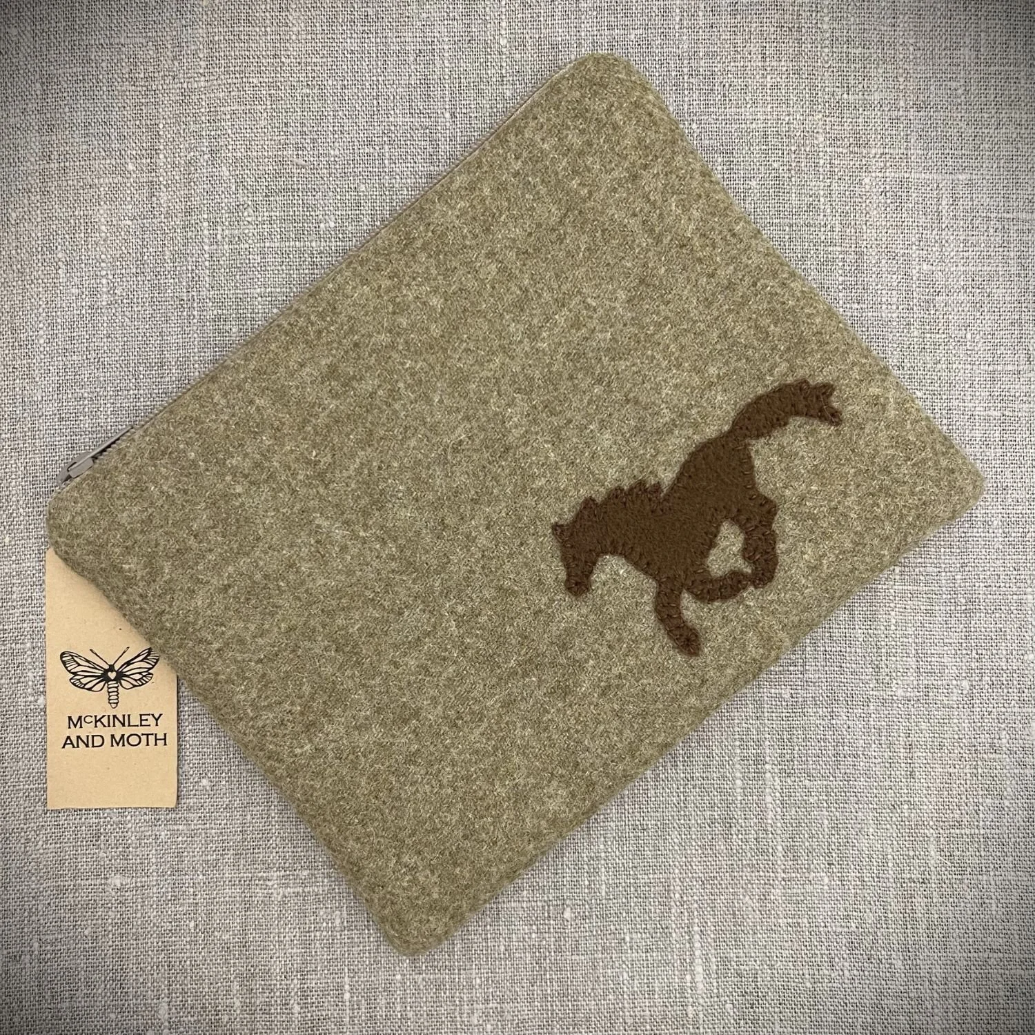 A small zipped bag made from a sandy coloured wool fabric with a small tan Horse silhouette stitched on the front.