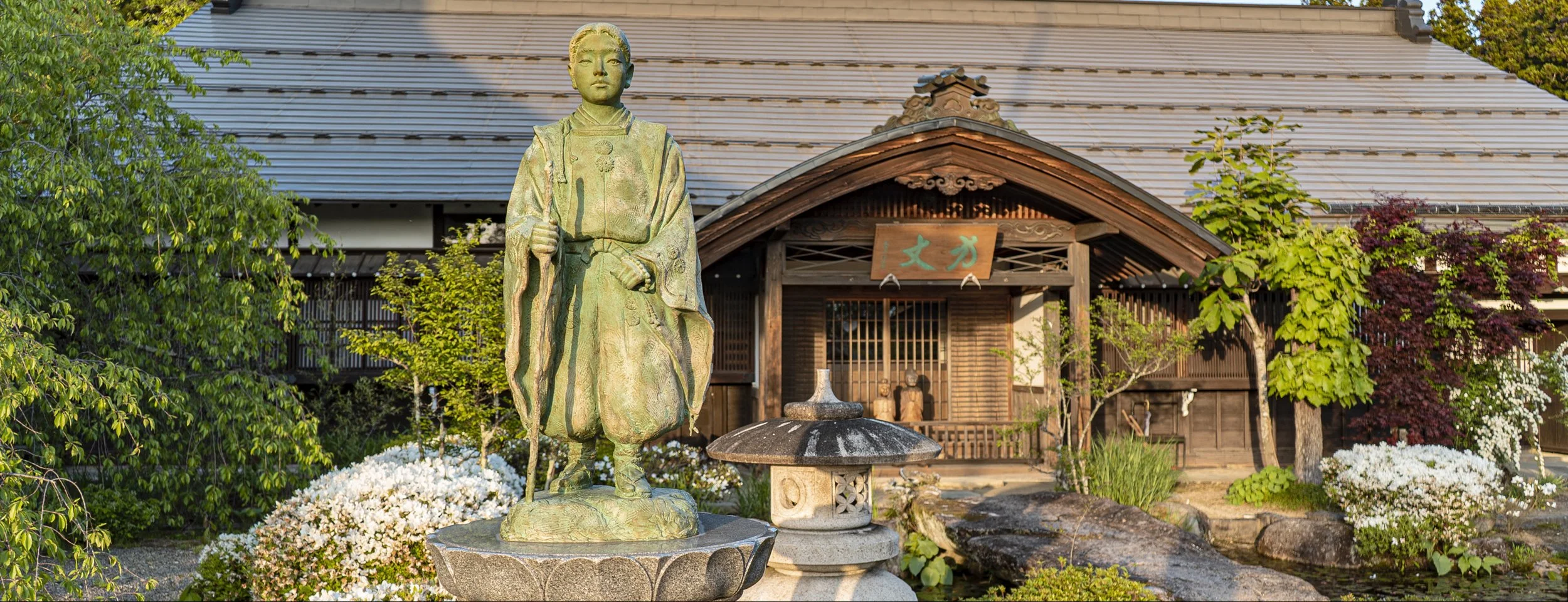 Bronze statue of a boy in traditional clothing standing in a garden in front of a Japanese-style wooden building with trees and flowers.