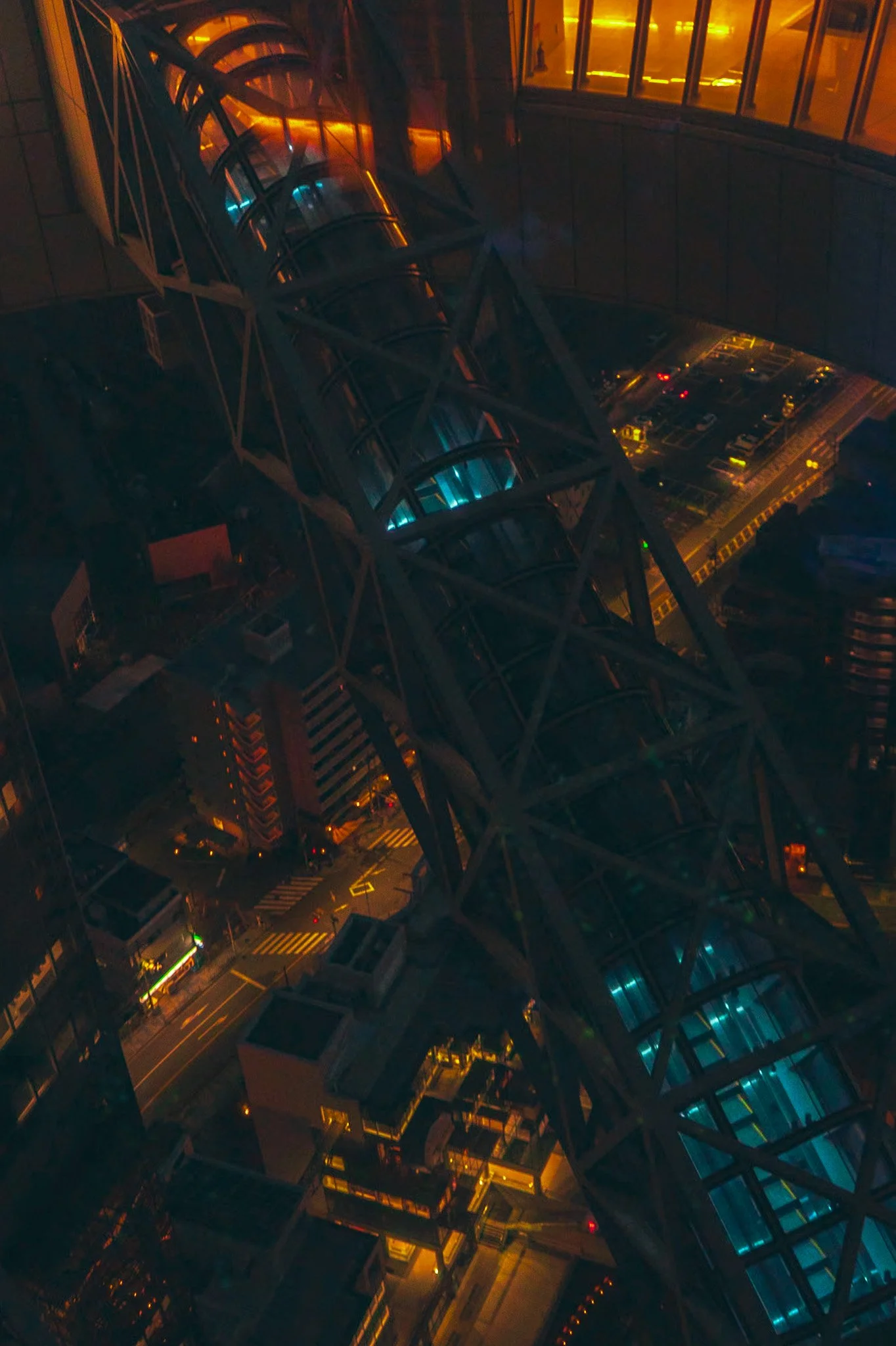 Nighttime view of a city street taken from a high level, featuring a modern, glass-enclosed walkway with blue lighting, surrounded by parking lots and lit buildings.