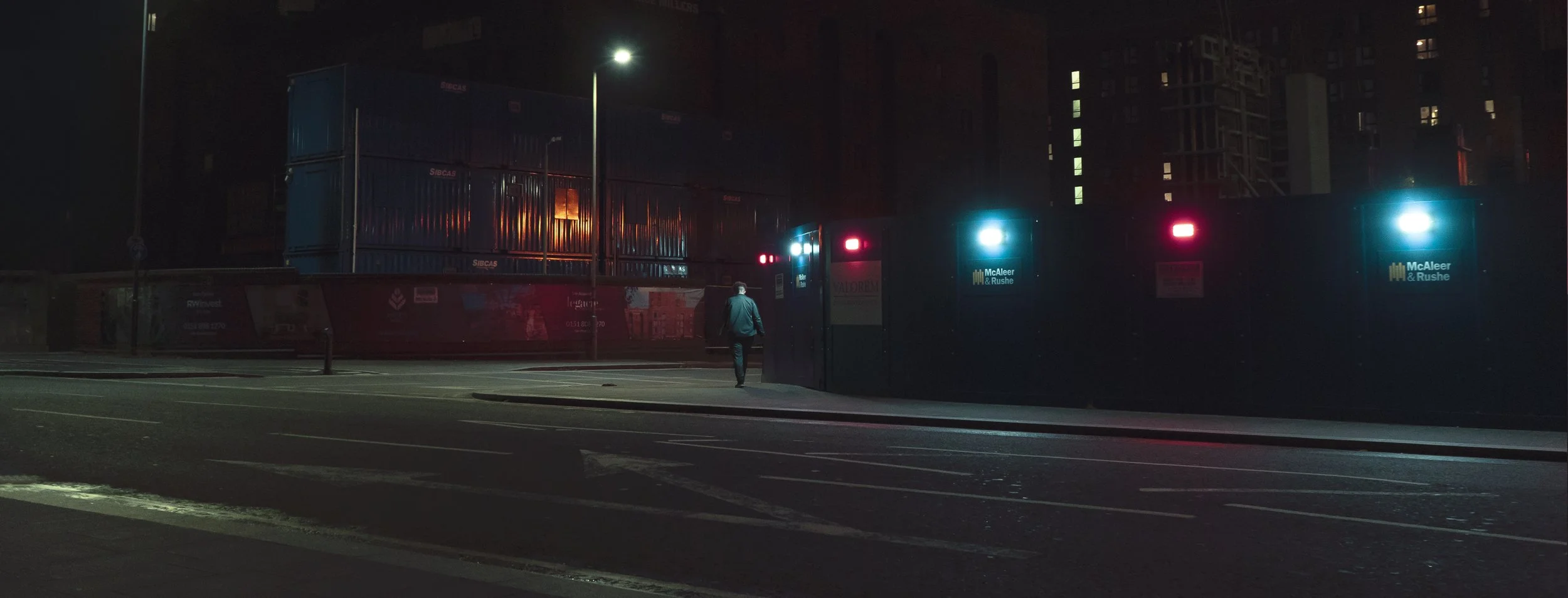 A man walking alone at night past construction site fencing with blue and red lights, with containers labeled SIBECAS, and illuminated buildings in the background.