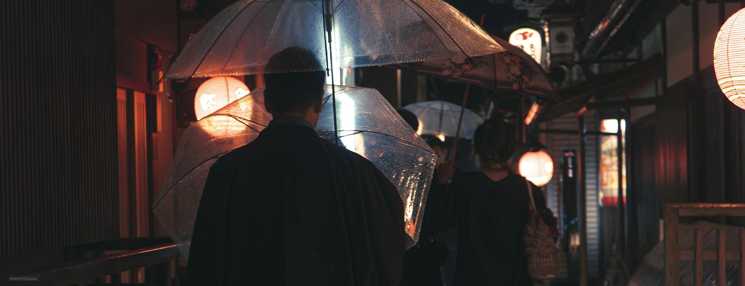 People walking in a rainy street at night with umbrellas illuminated by streetlights and lanterns.