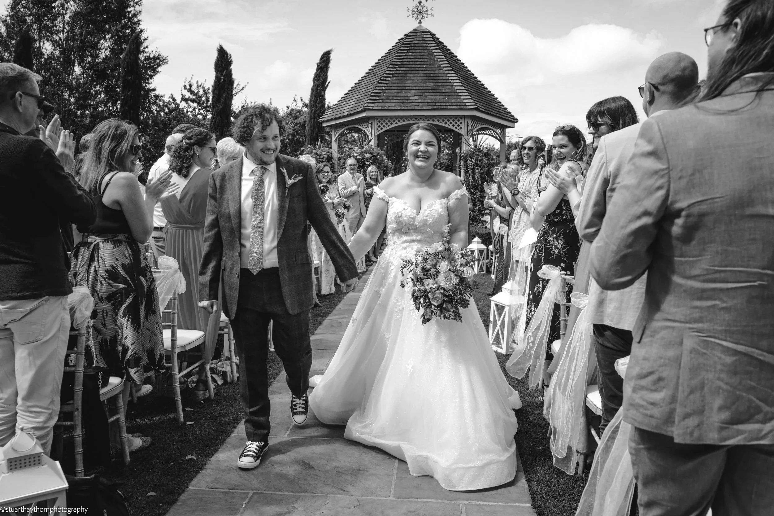 A bride and groom walking down the aisle, smiling and holding hands, surrounded by guests clapping during a wedding ceremony outdoors. The bride is wearing a wedding dress and holding a bouquet, while the groom is in a suit. There is a gazebo in the background, and people are dressed in semi-formal attire.