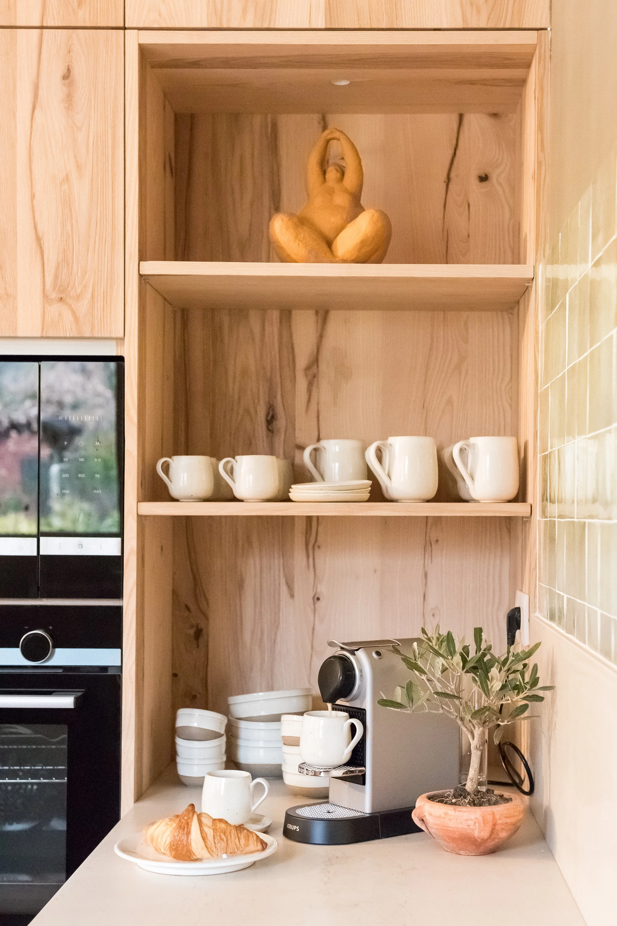 Kitchen shelf with a statuette, white cups and plates, coffee machine, and a potted plant with a croissant and coffee mug on the counter.