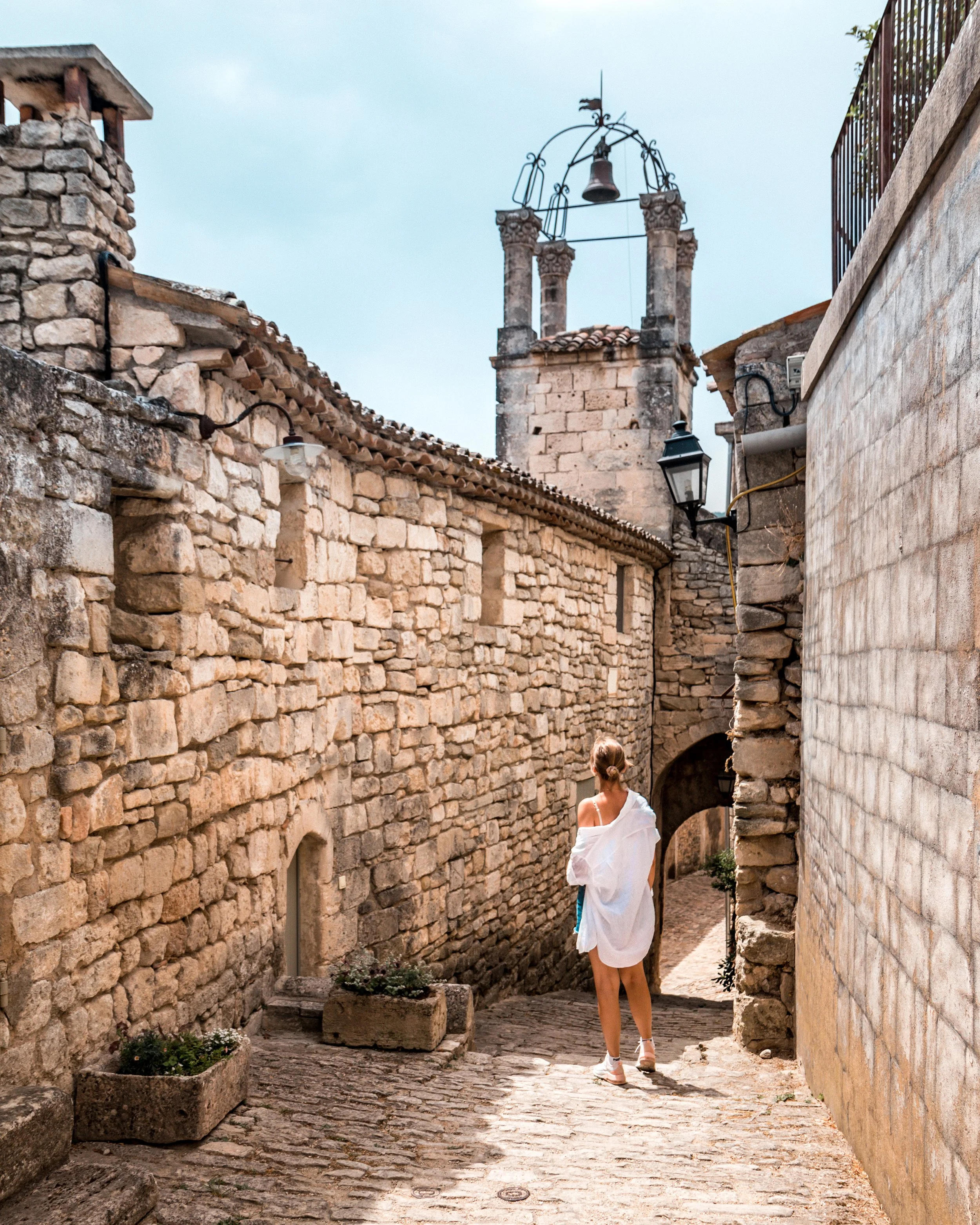 Woman standing on a cobblestone alleyway with stone buildings on both sides and a bell tower in the background.