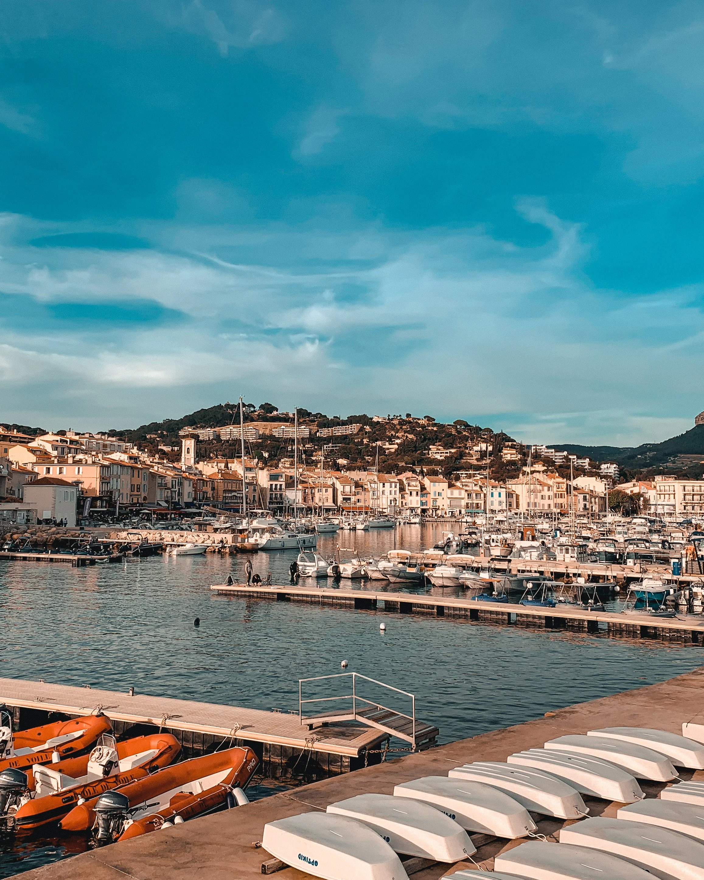 View of a marina with boats docked, colorful residential buildings along the waterfront, hills in the background, and a blue sky with some clouds.