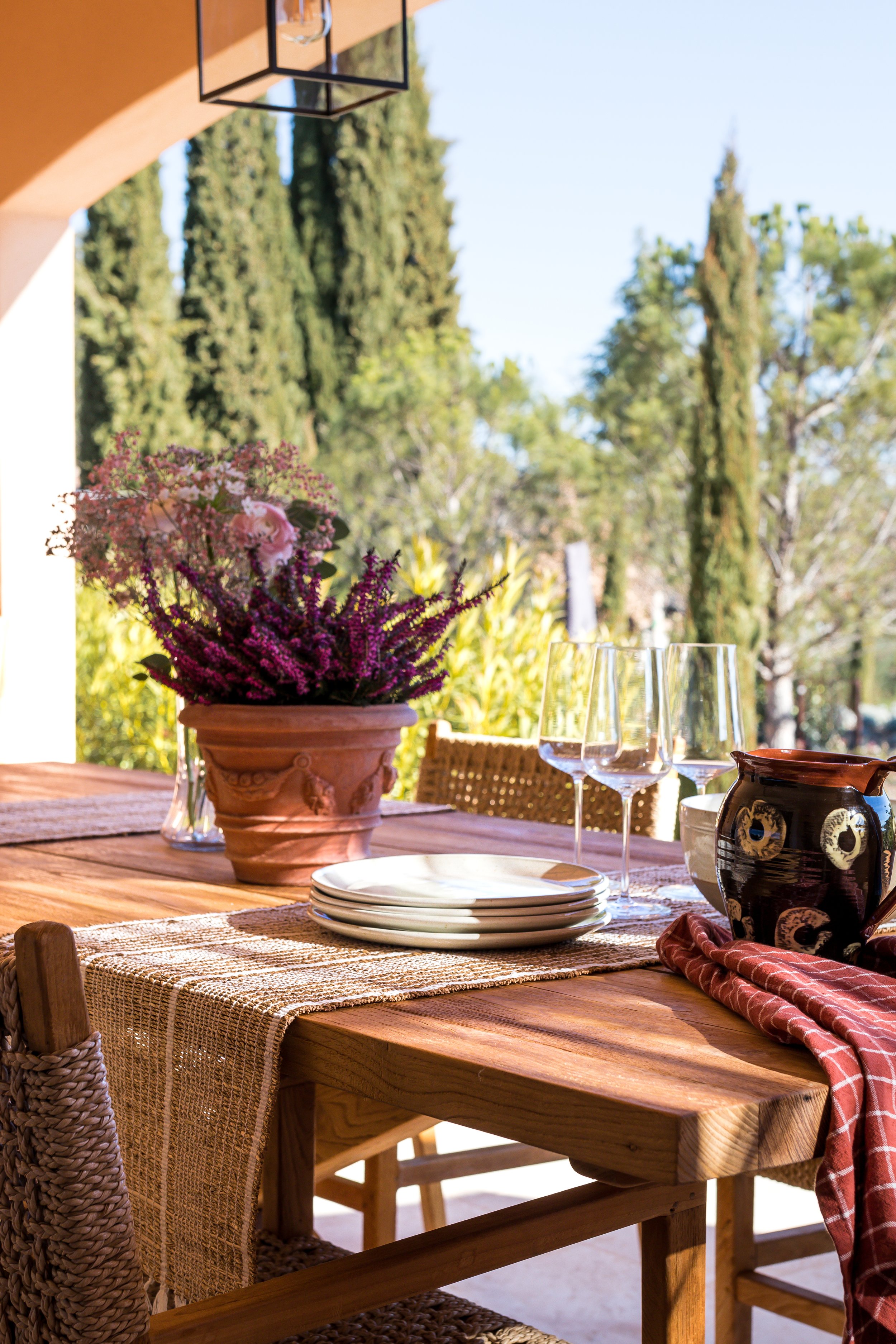 An outdoor dining table set with a flower arrangement, plates, wine glasses, a decorative pot, and a cloth napkin, with lush greenery in the background.