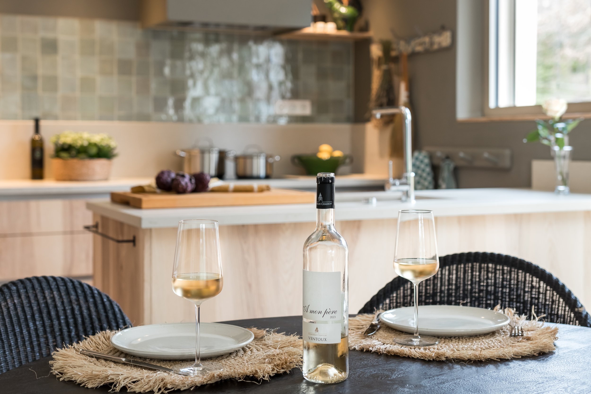 A dining table set with two plates, two wine glasses filled with white wine, and a bottle of wine. The table has woven placemats and is situated in a modern kitchen with a sink, window, and various kitchen items in the background.