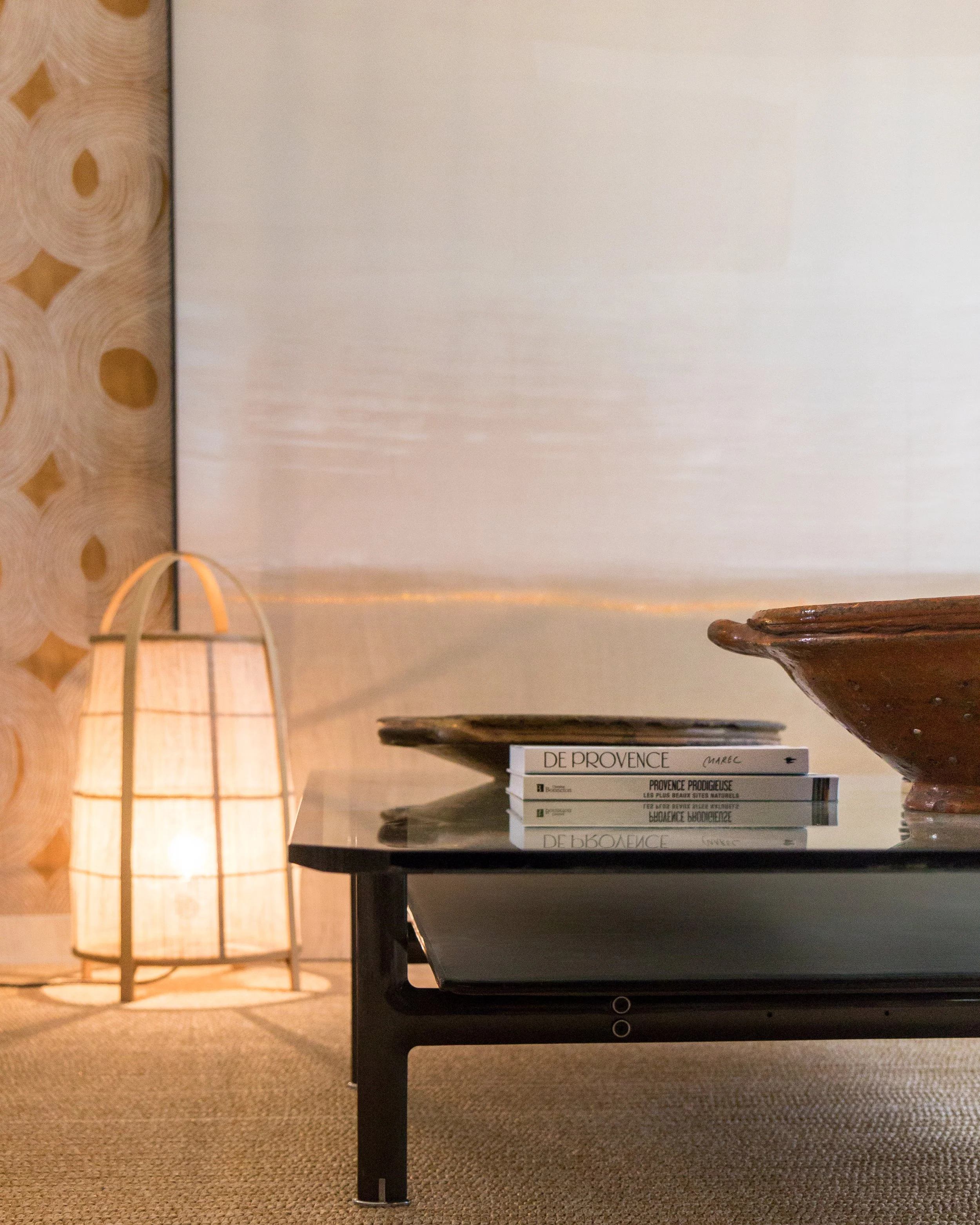A modern interior scene featuring a black table with a glossy top holding a stack of books, a decorative pottery dish, a warm floor lamp with a paper shade, and a textured wallpaper with circular patterns in the background.