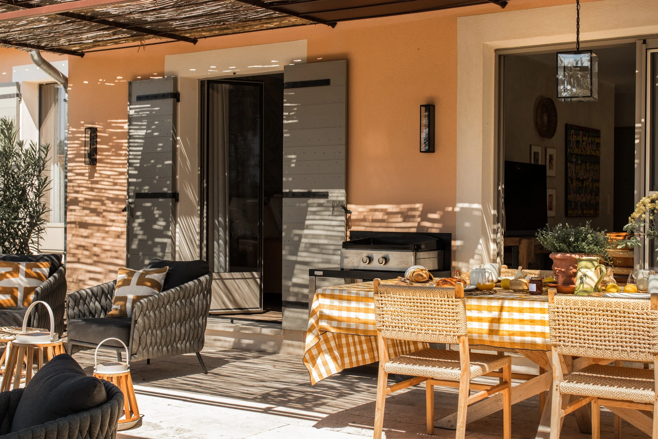 Outdoor patio with a table covered in a yellow and white checkered tablecloth, surrounded by wicker and upholstered chairs. There are outdoor living furnishings, potted plants, and a barbecue grill under a partial roof with sunlight casting shadows.