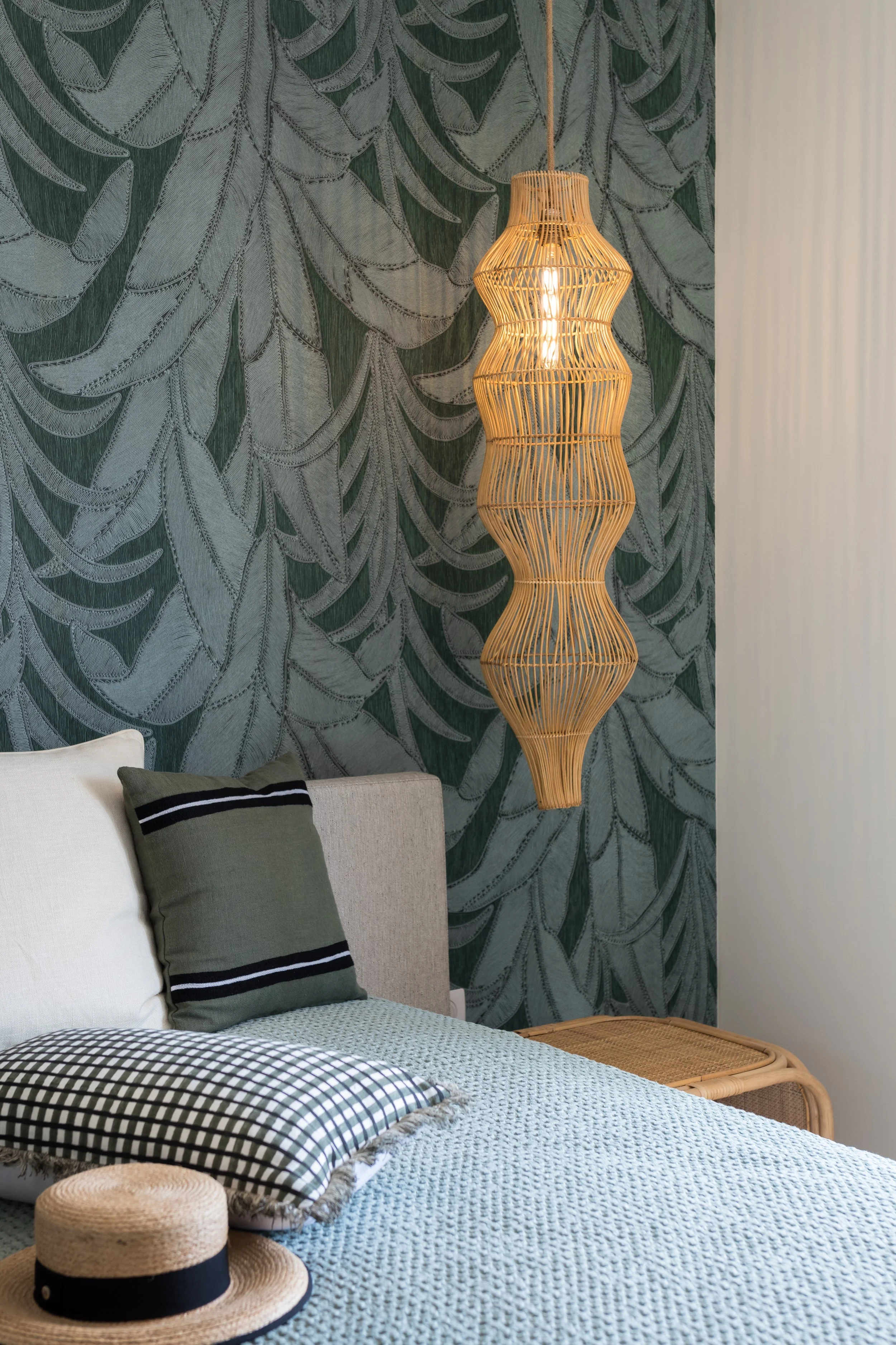 Interior of a bedroom featuring a bed with pillows and a woven hat, a rattan hanging light fixture, and a wall with leafy patterned wallpaper.