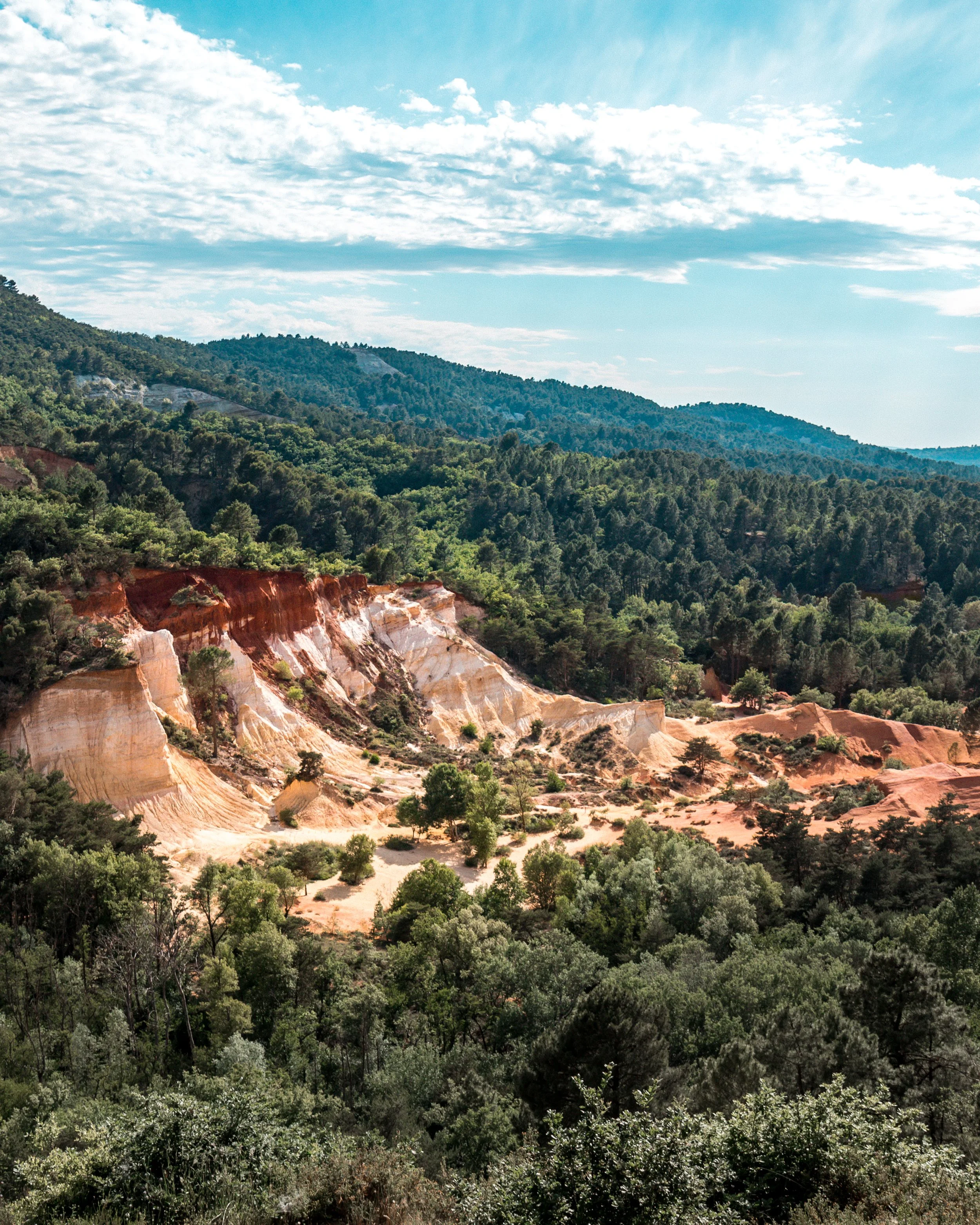A scenic view of a forested mountain landscape with colorful, layered cliffs and a partly cloudy sky.