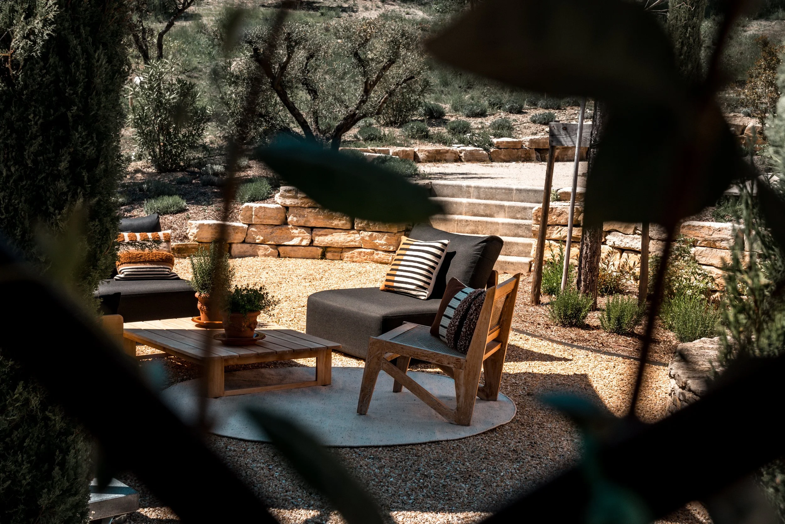 A cozy outdoor seating area with a black lounge chair, striped cushions, a wooden chair, a small coffee table with potted plants, and a stone staircase with rock wall in the background, viewed through dense foliage.