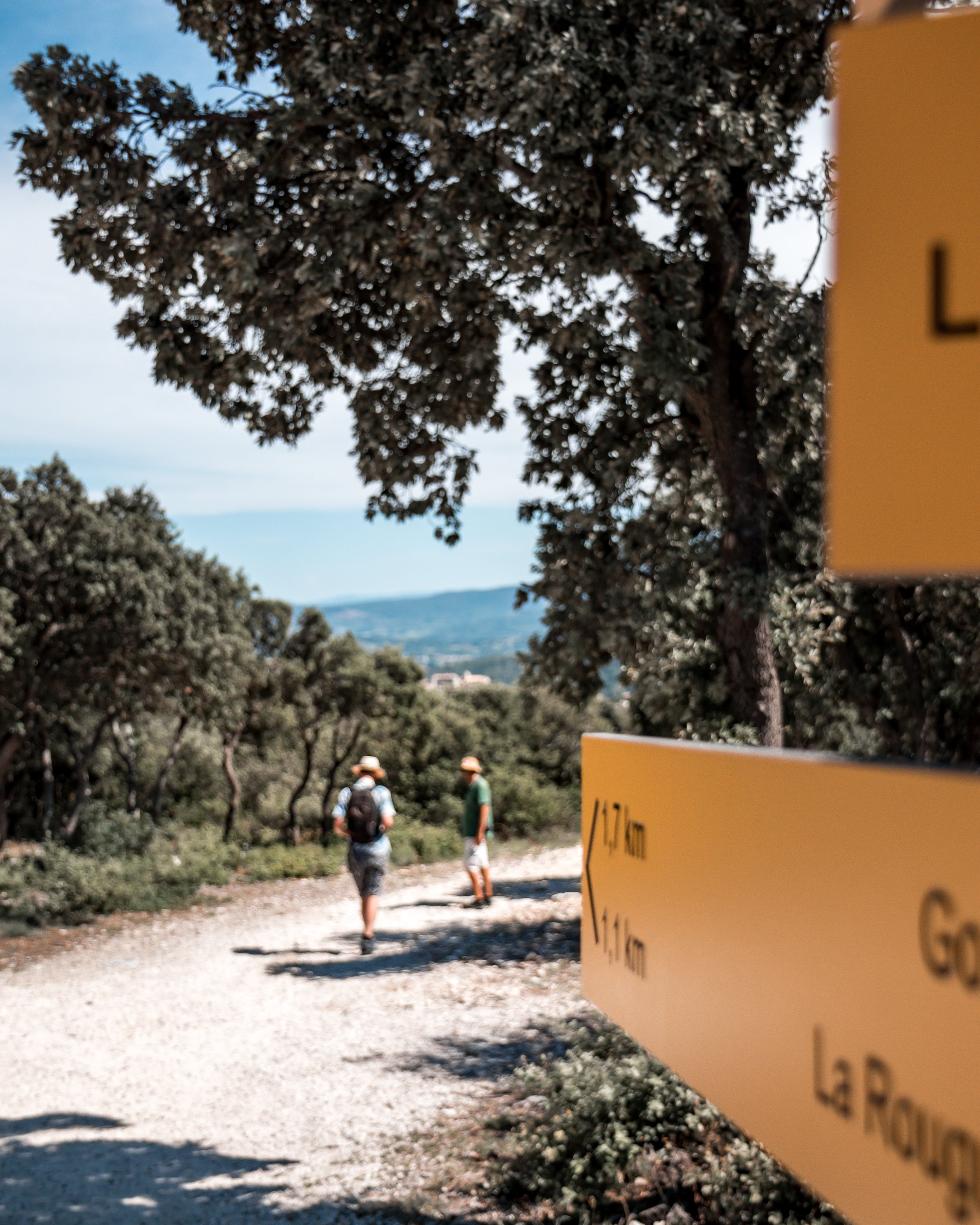 Hikers walking on a gravel trail in a wooded area with trees and a distant view of mountains in the background. A yellow sign with distance information is partially visible in the foreground.