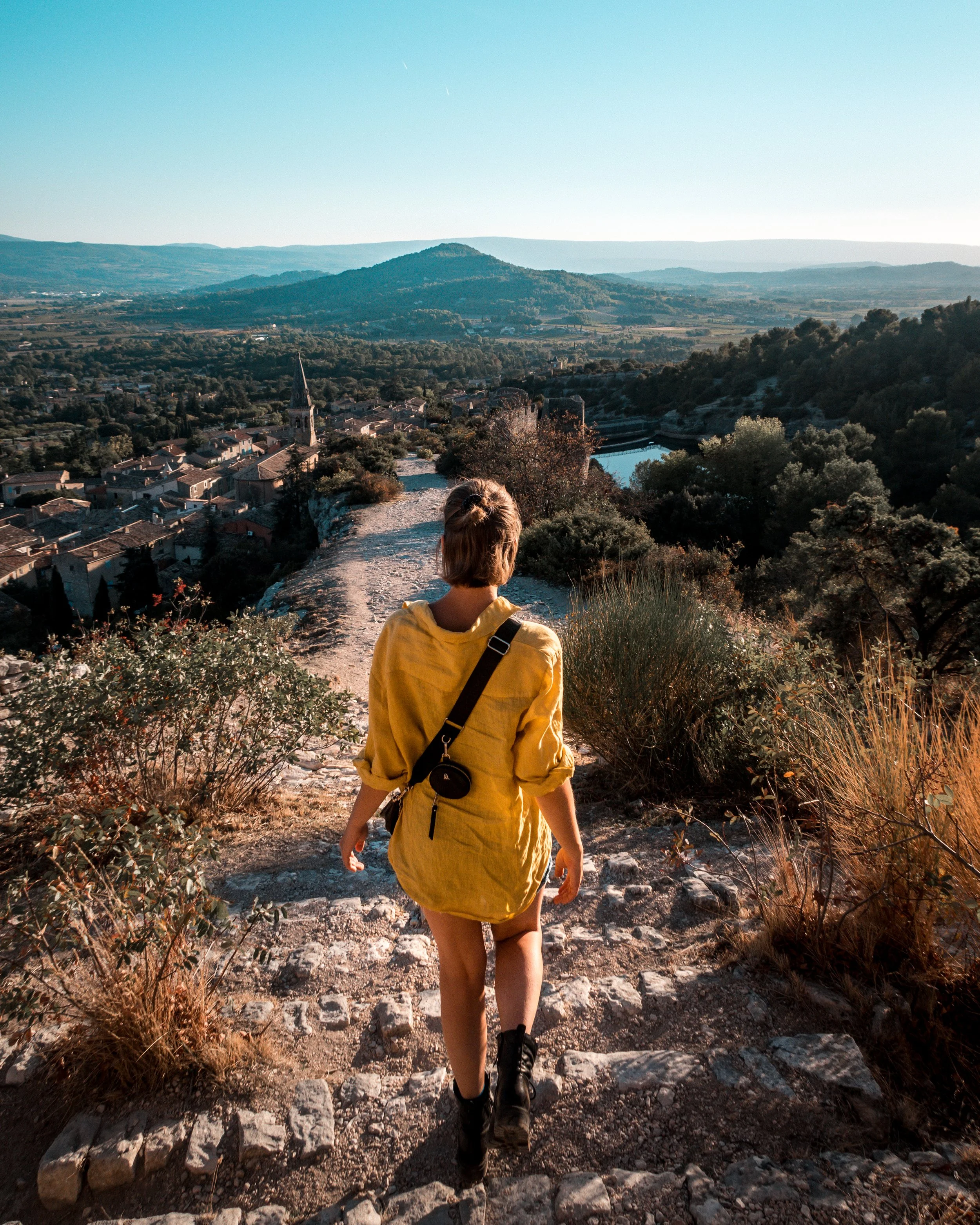 A woman in a yellow jacket and black boots hiking on a rocky trail overlooking a small town with a church steeple, surrounded by green trees and distant hills.