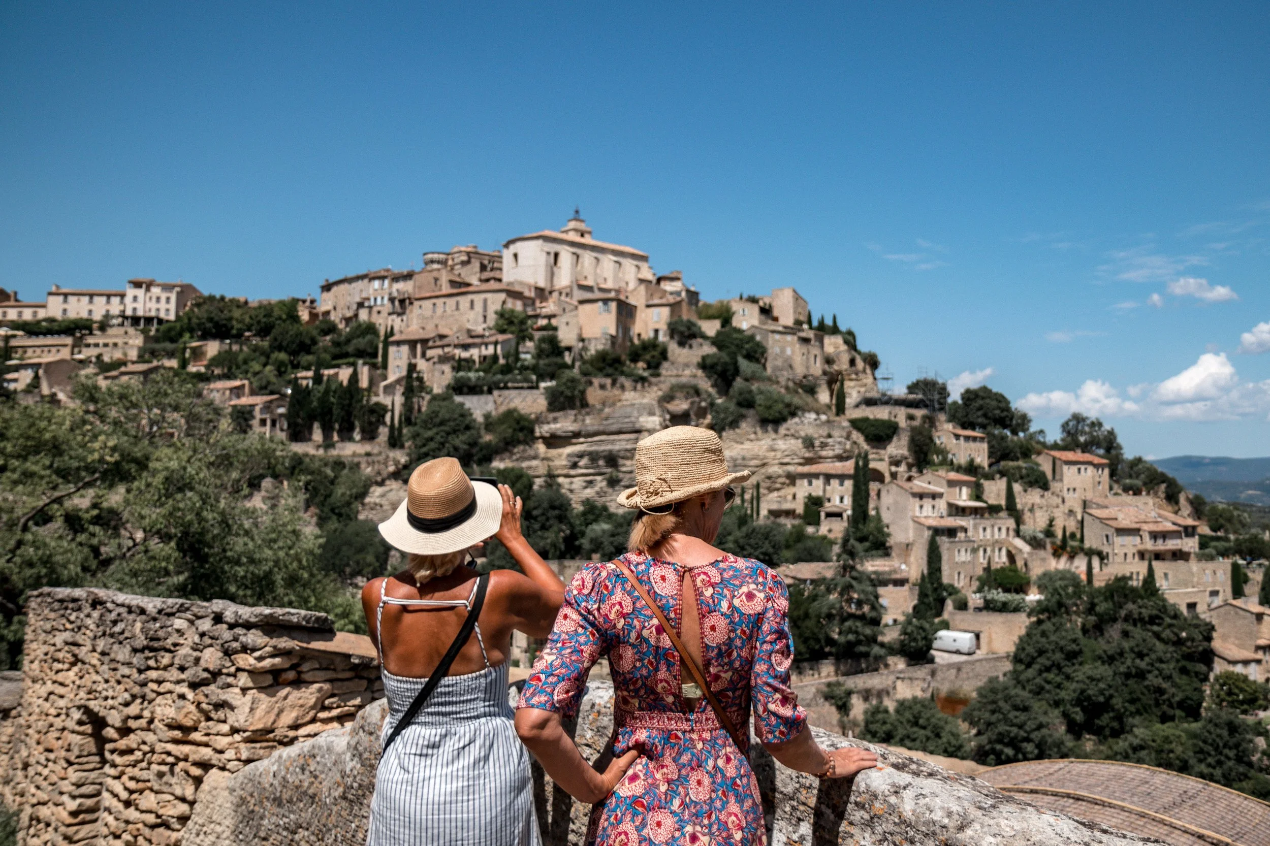 Two women wearing sun hats standing on a stone balcony overlooking a hillside town with old buildings and lush greenery under a bright blue sky.
