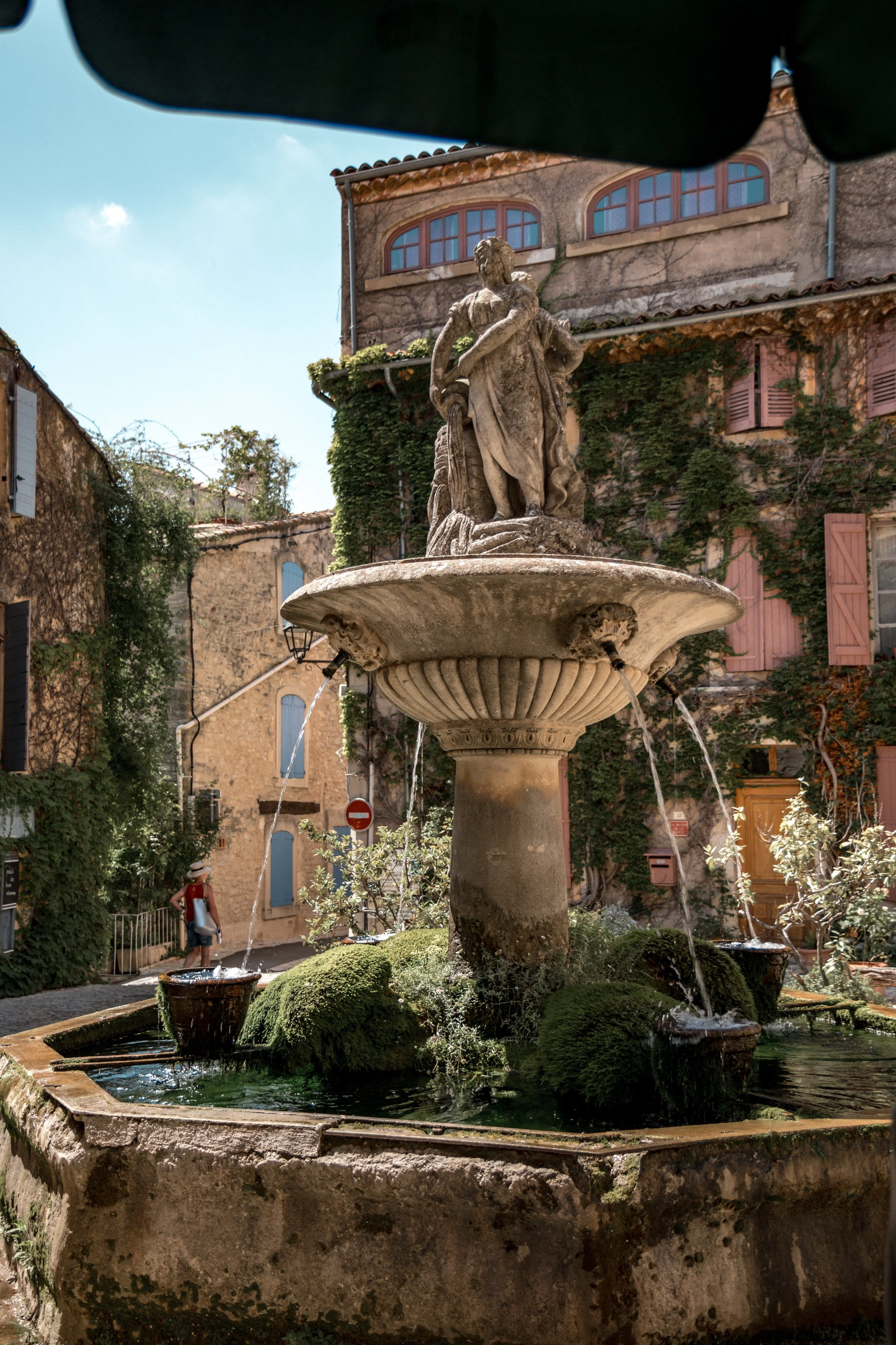 Stone fountain with a sculpture of a woman standing in the center, water flowing from four spouts into a basin, surrounded by greenery and old buildings in the background.