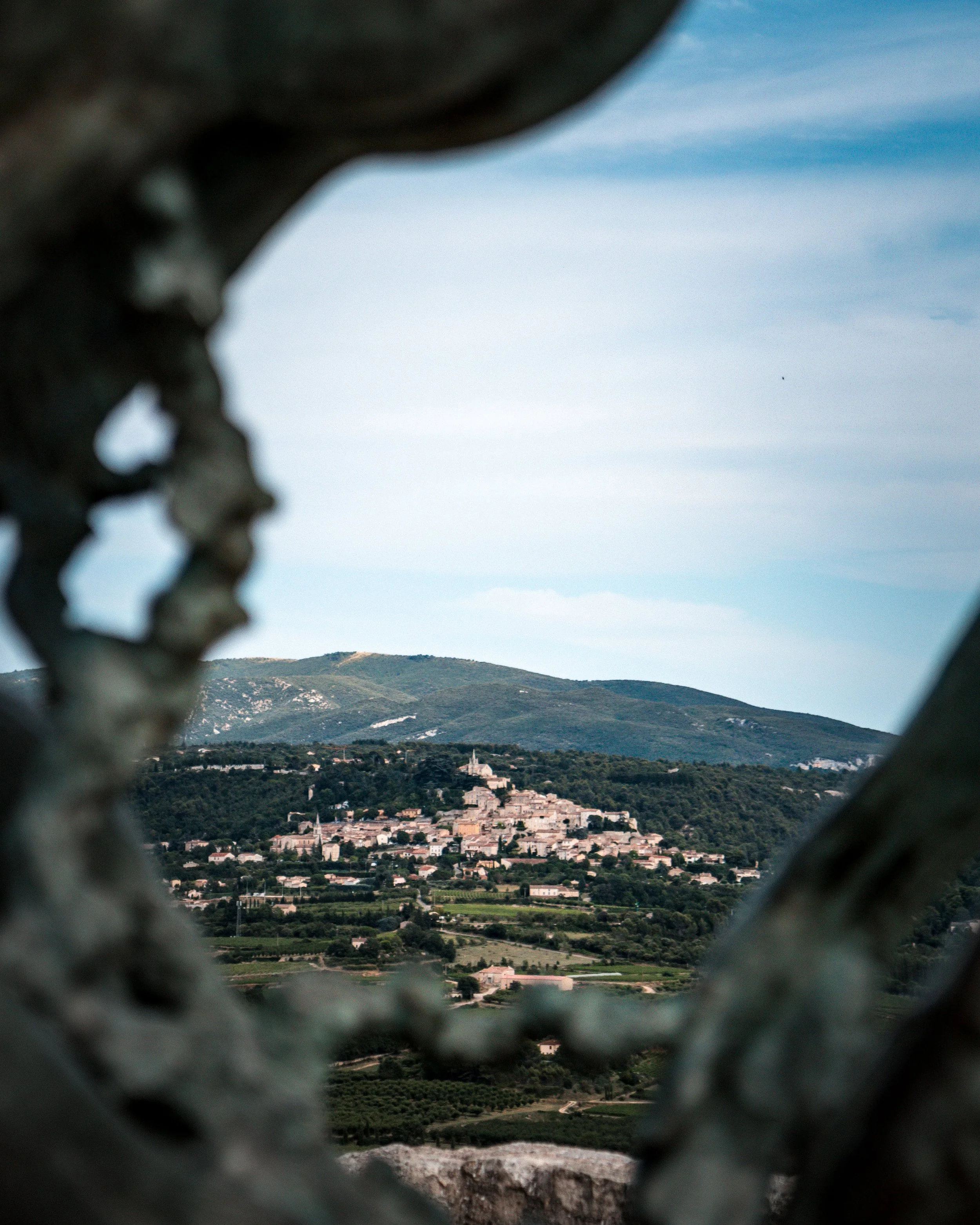 View of a hilltop town through a decorative metal lattice, with green fields and mountains in the background.
