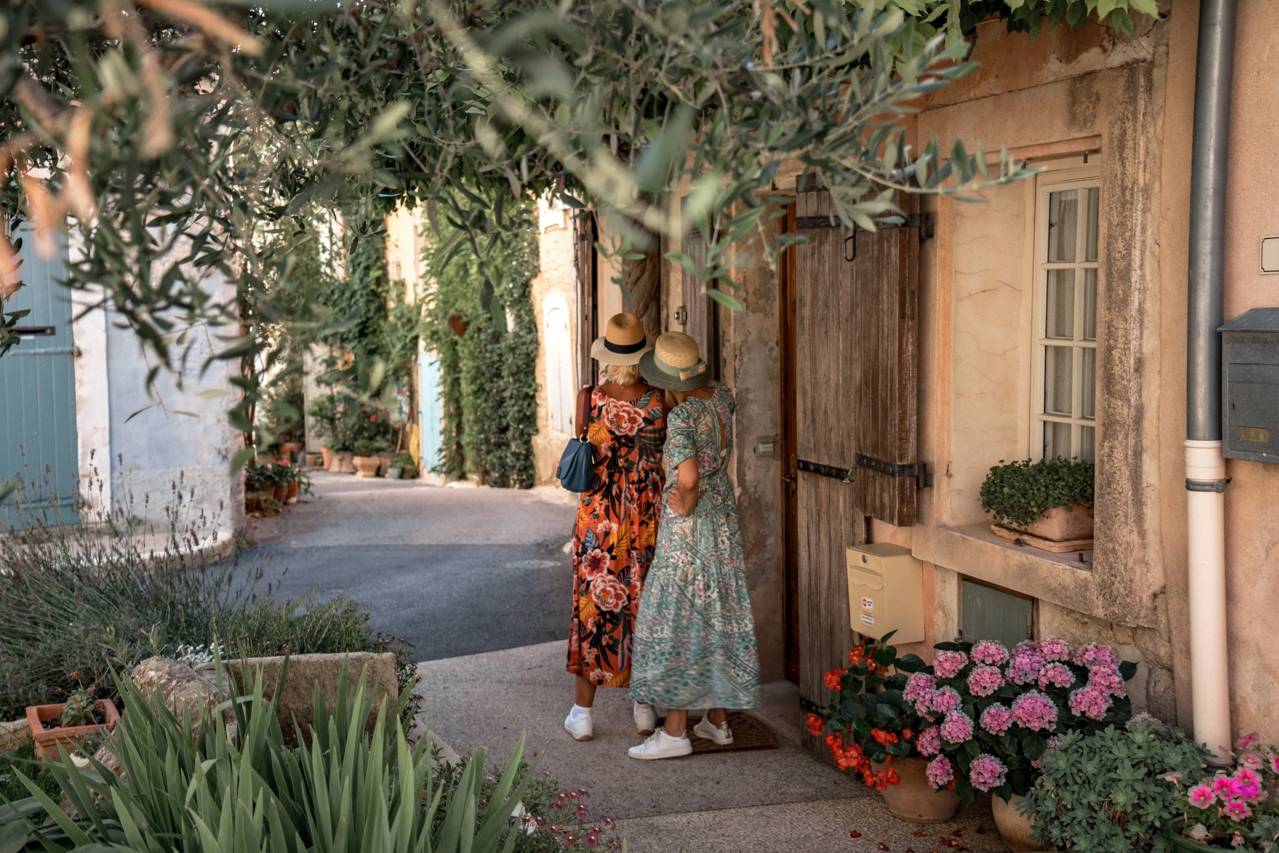 Two women in colorful dresses and wide-brimmed hats standing outside a rustic house with potted plants and flowers, talking to each other on a narrow street.