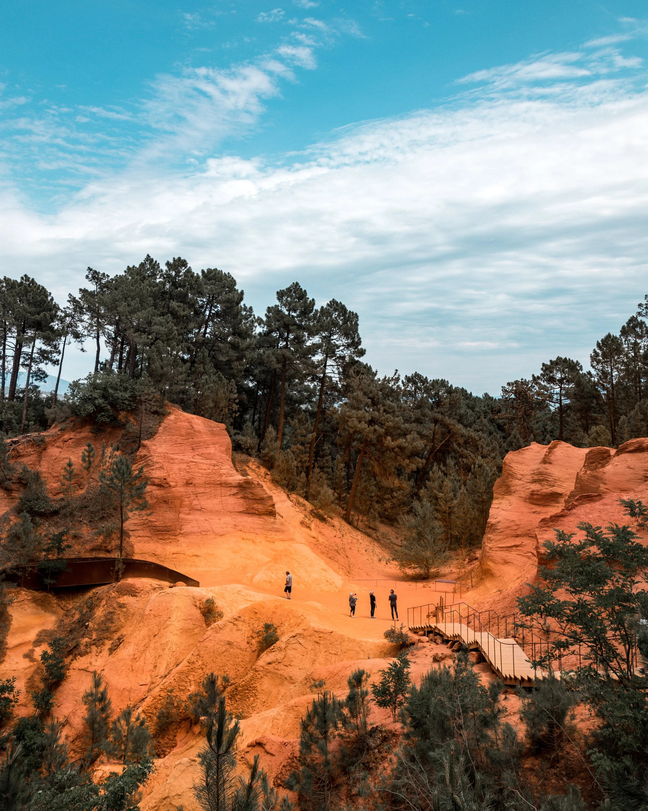 People walking on a trail through red sandstone formations surrounded by pine trees under a cloudy blue sky.