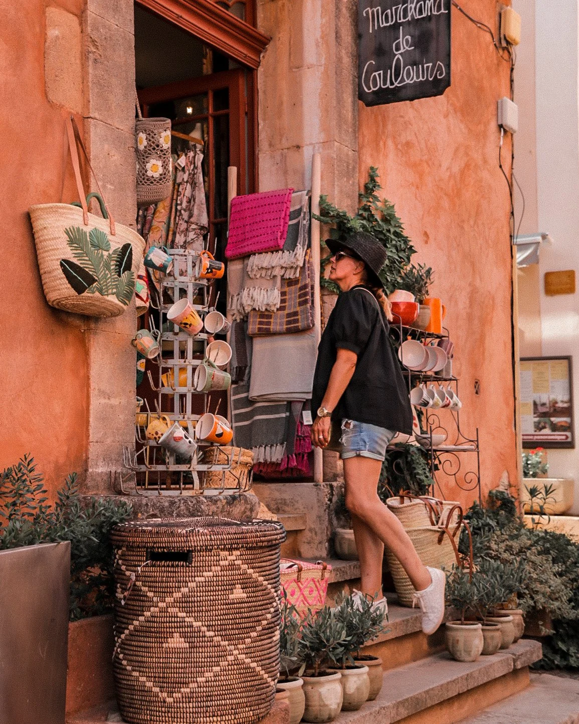 A woman in a black wide-brimmed hat, black top, and denim shorts browsing souvenirs outside a colorful shop with orange walls. The shop display includes handbags, cups, blankets, and potted plants.