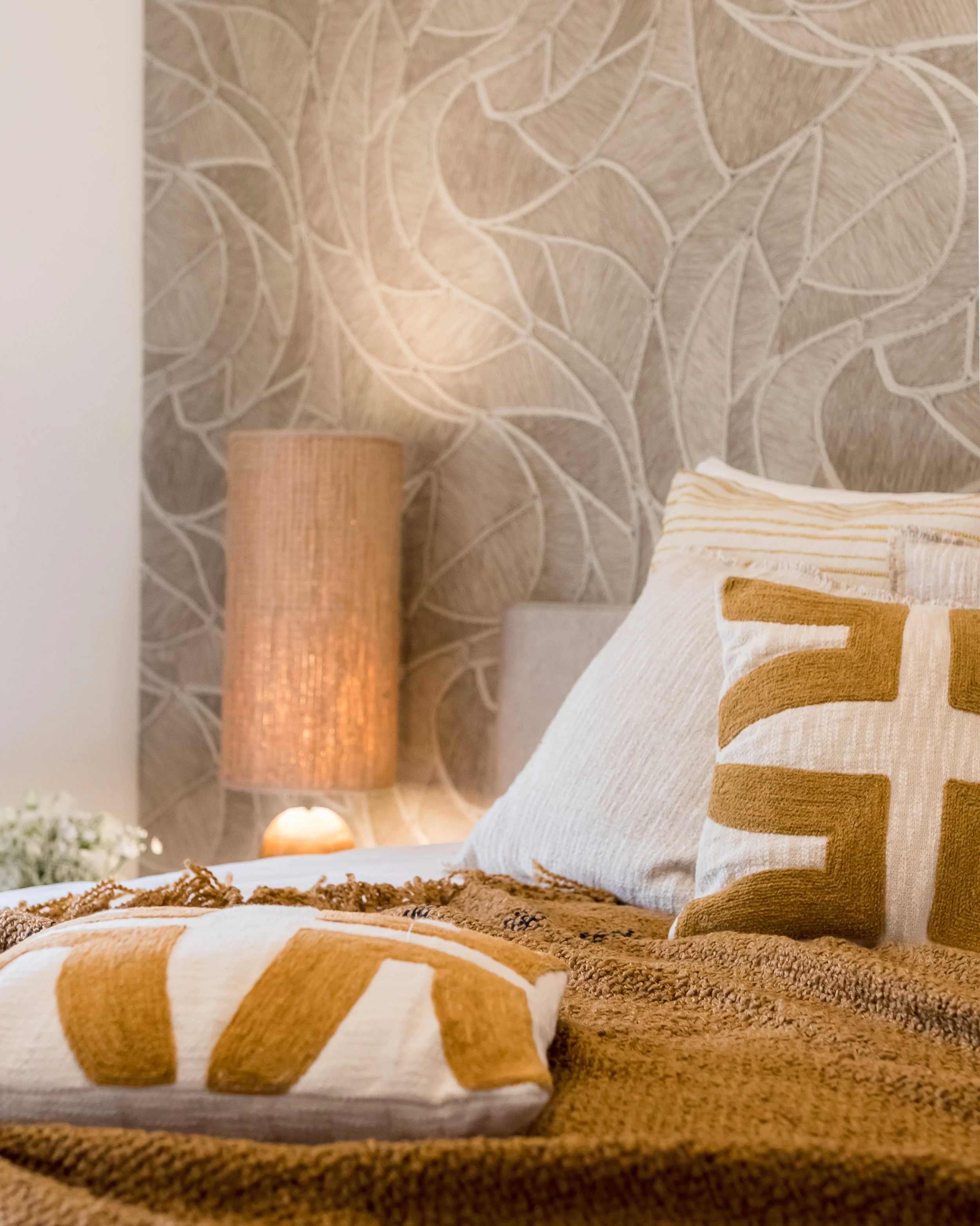 A cozy bedroom with a beige upholstered headboard, decorative throw pillows with beige and gold geometric patterns, a warm brown textured blanket, a woven nightstand with an orange lamp, and patterned wallpaper with abstract leaf designs.