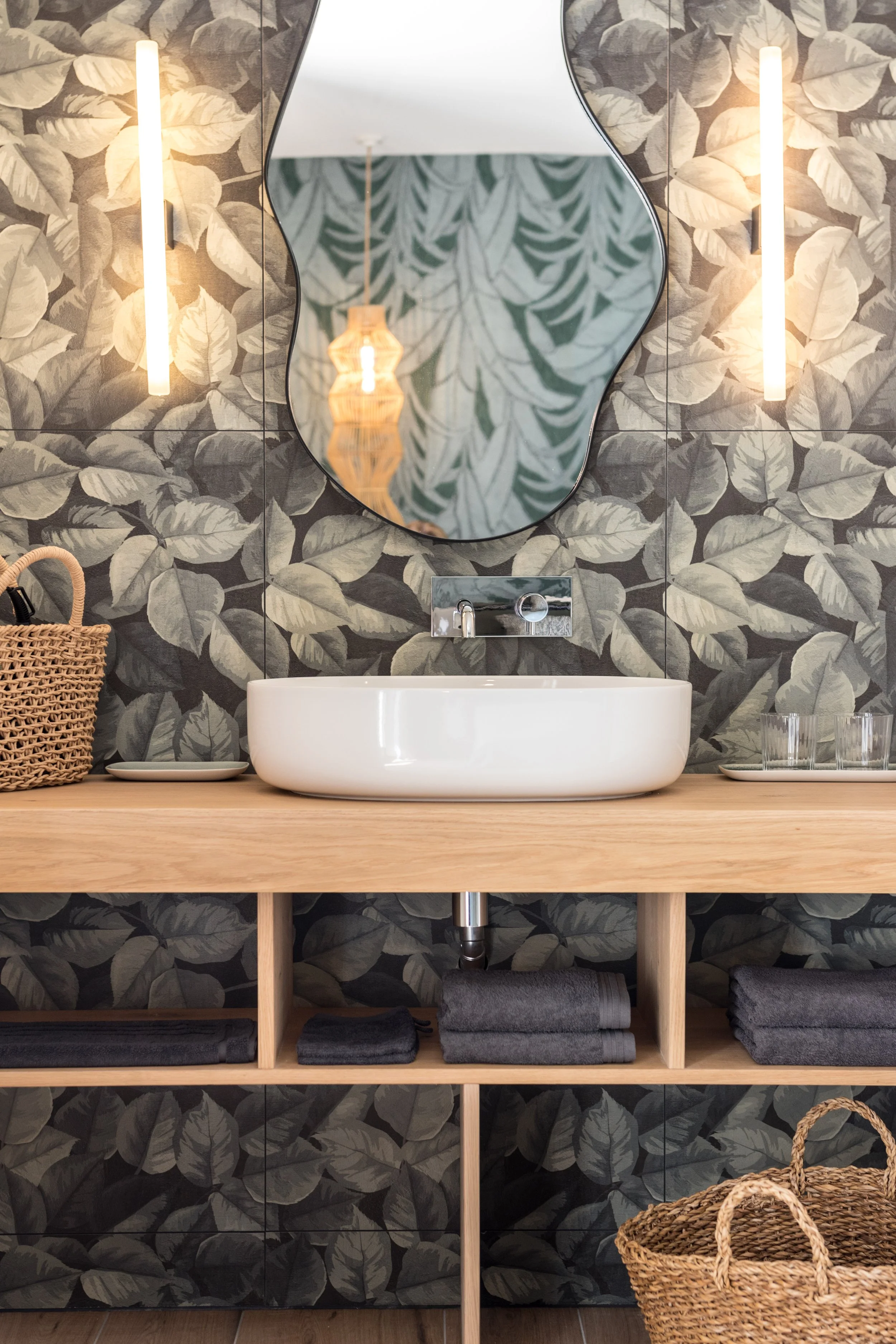 A bathroom vanity with a white vessel sink, a large irregularly shaped mirror, and leafy patterned wallpaper. The vanity has a wooden countertop with shelves holding gray towels and a woven basket. Two wall-mounted lights are on either side of the mirror, and a reflection of a hanging pendant light can be seen in the mirror.