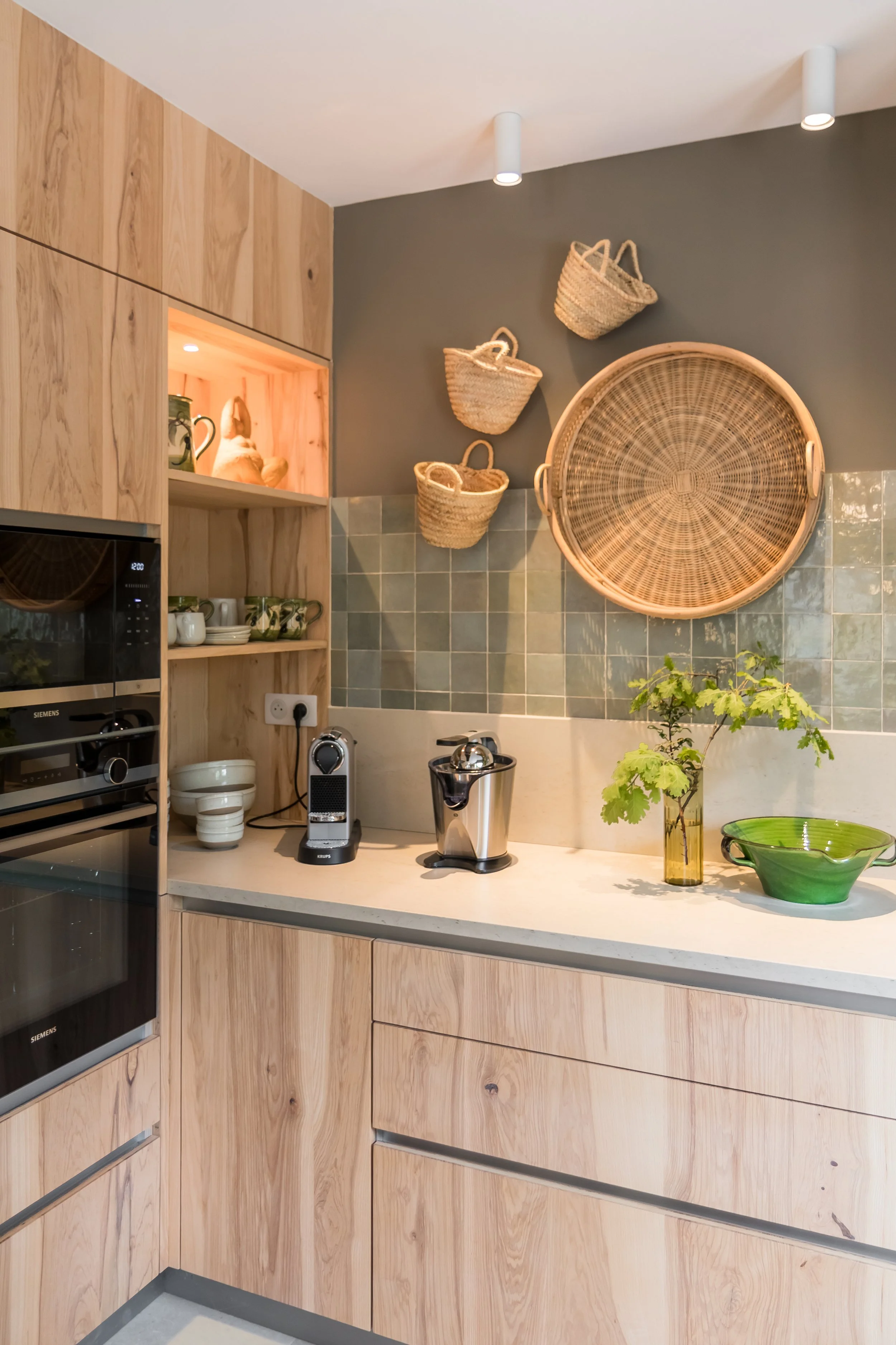 A kitchen with light wood cabinets, a white countertop, and a tiled backsplash. Decor includes woven baskets on the wall, green ceramics, and a small plant in a glass vase.