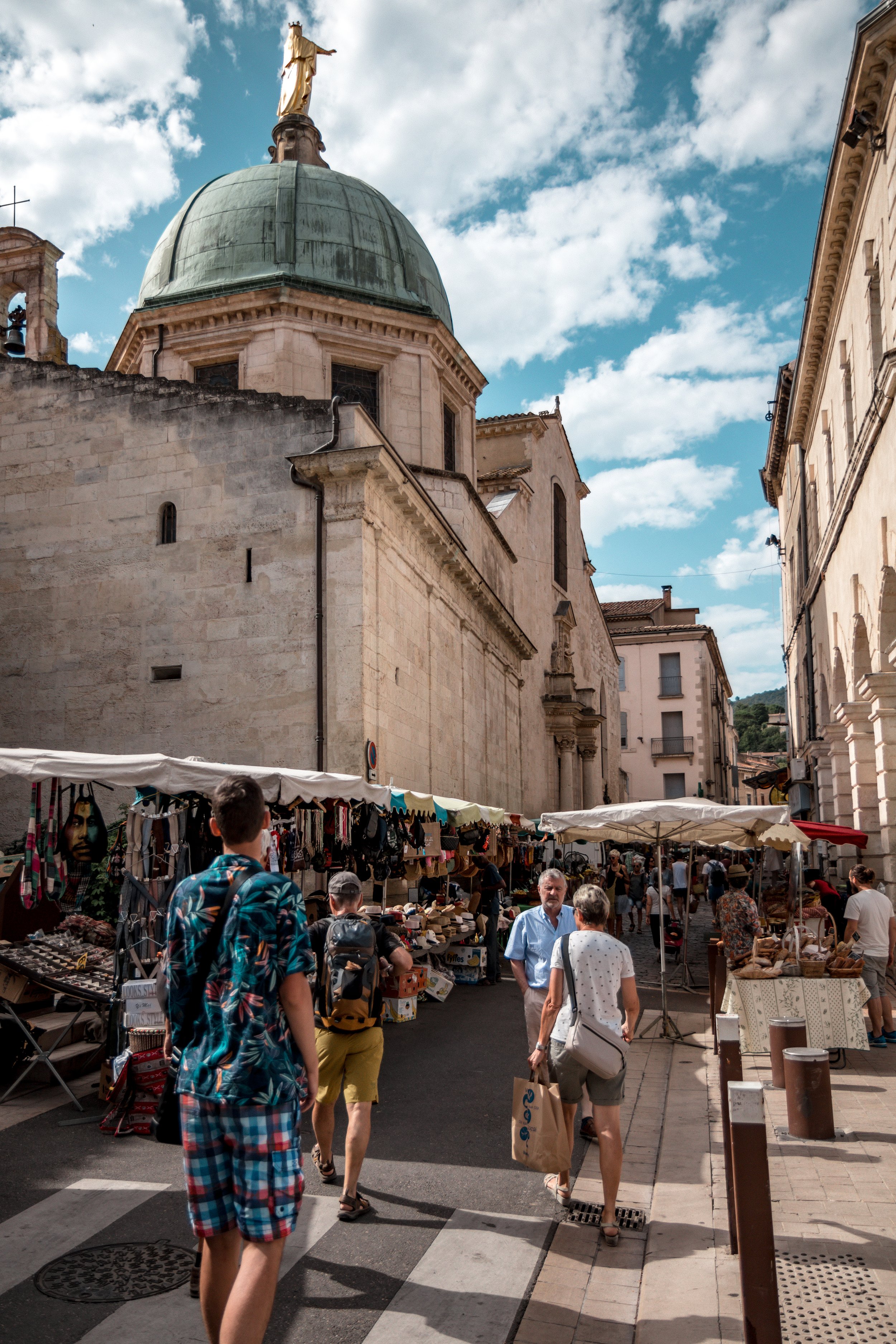 People walking through an outdoor market on a city street with historic buildings and a church with a large green dome and a statue on top underneath a partly cloudy sky.